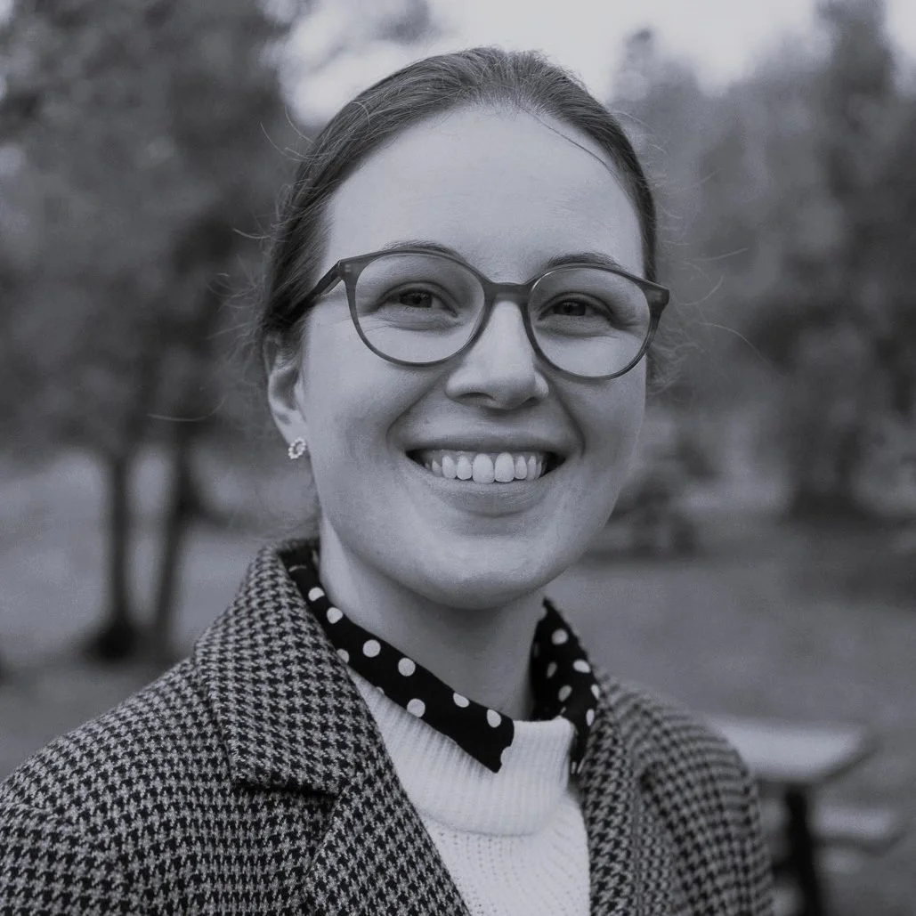 Black and white photo of Cathleen Mair wearing a tweed jacket and white jumper and round glasses with a park in the background