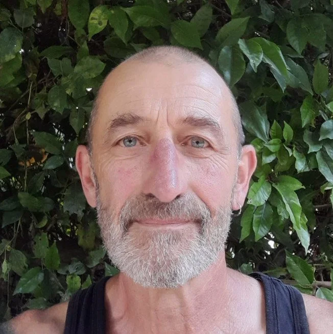 Photo of Alan Hughes looking face on wearing a tank top standing in front of a tall green bush