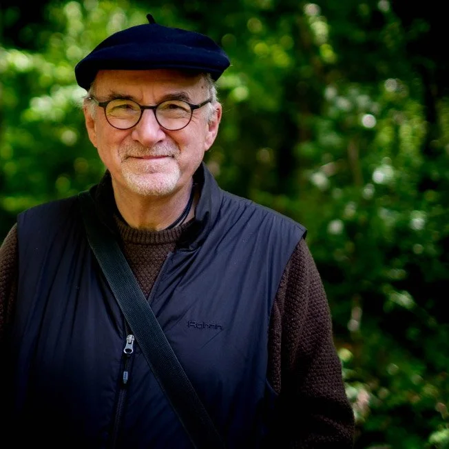 Photo of Philip Rush wearing a cap, brown jumper and black Gillet, standing with trees in the background
