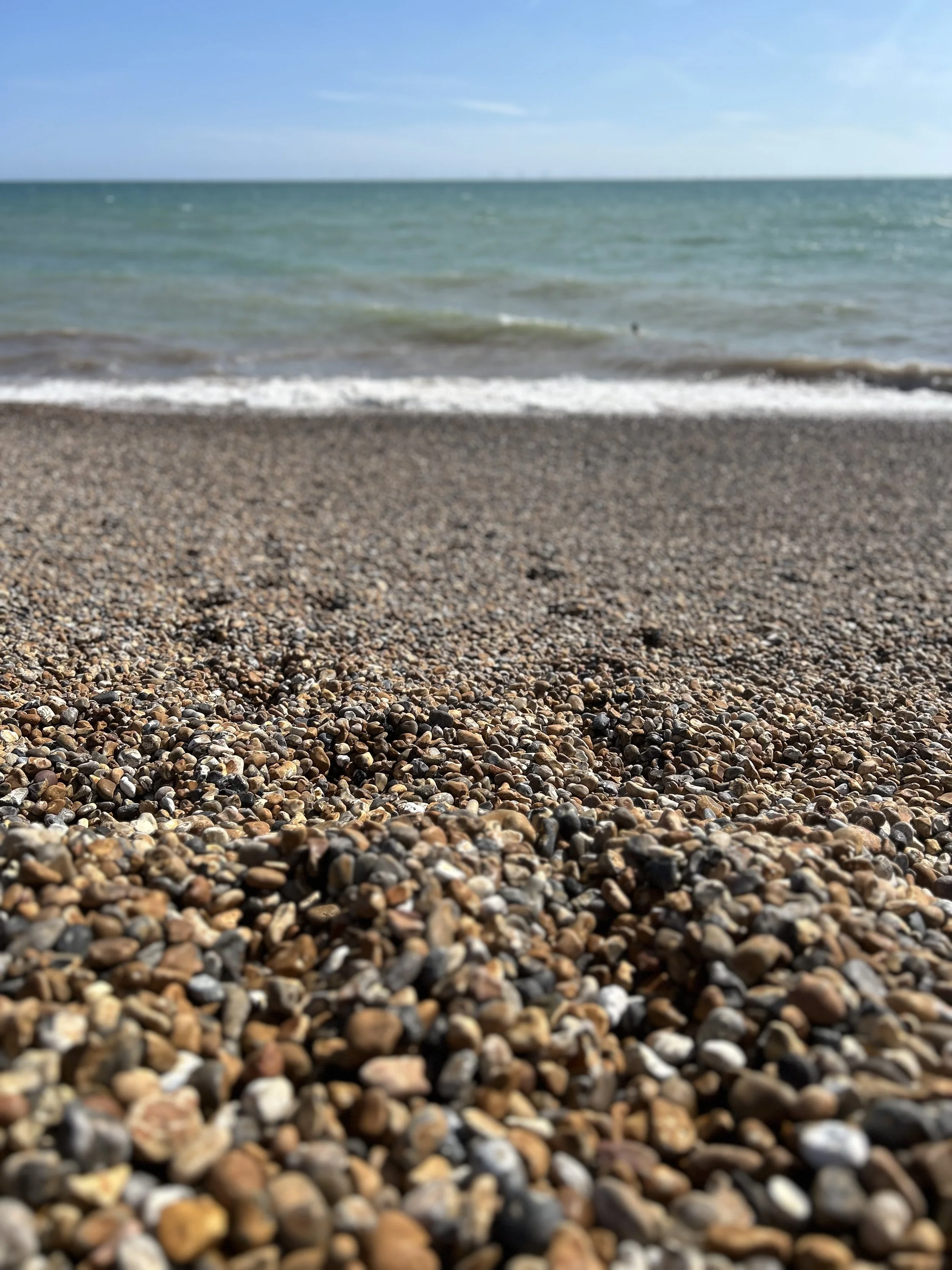 Close-up view of a pebble beach with small, multicoloured pebbles in the foreground and the ocean waves gently rolling in the background under a clear blue sky.