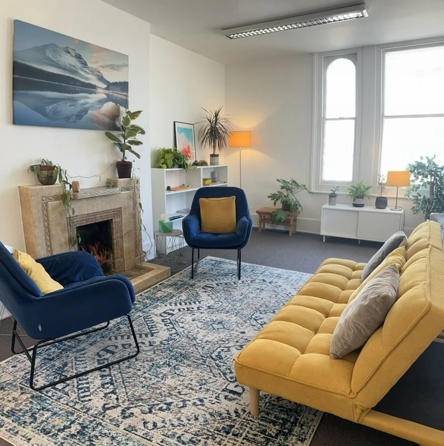 Bright living room with a yellow sofa, blue armchairs, a white fireplace, and large windows with natural light, decorated with plants, lamps, and wall art.