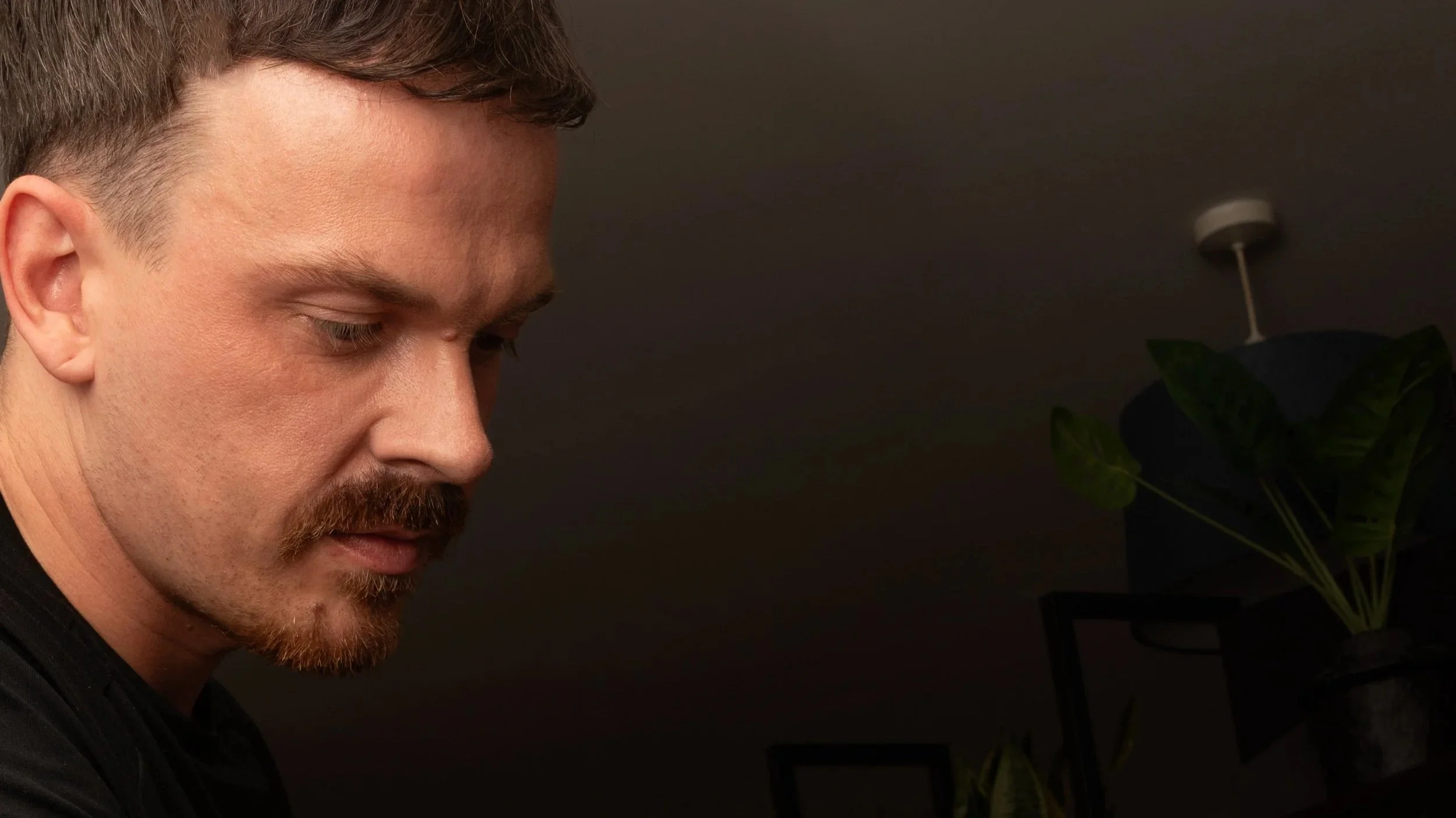 Close-up of a man with short brown hair, a mustache, and a beard, looking downward with a serious expression, against a dark background with a plant on the right.