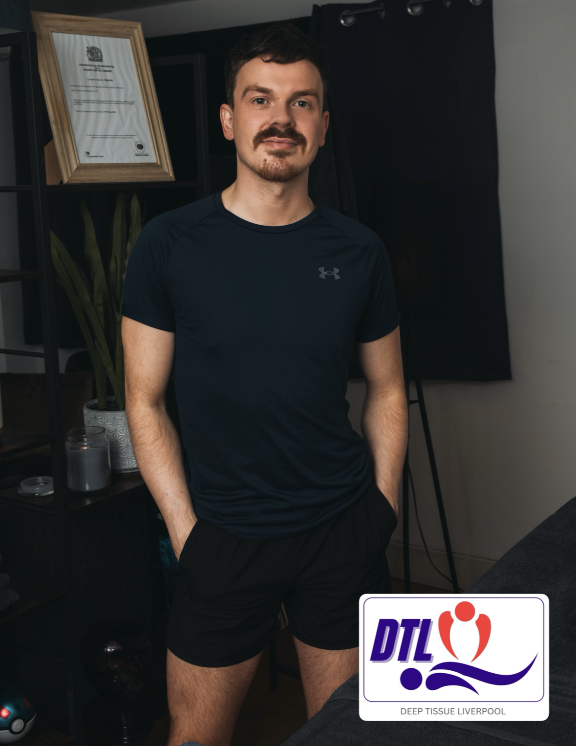 A young man with dark hair and a beard, wearing a black Under Armour athletic shirt and black shorts, standing indoors with his hands in his pockets. There is a framed certificate or diploma on the wall behind him, a potted plant, and a black curtain or backdrop. A logo for "DTL" and the words "Deep Tissue Liverpool" are visible in the bottom right corner of the image.