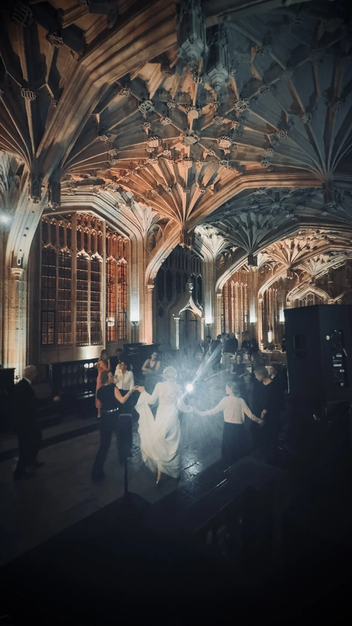 People dancing in a grand hall with high, ornate vaulted ceiling and large, stained glass windows, illuminated by warm lighting.