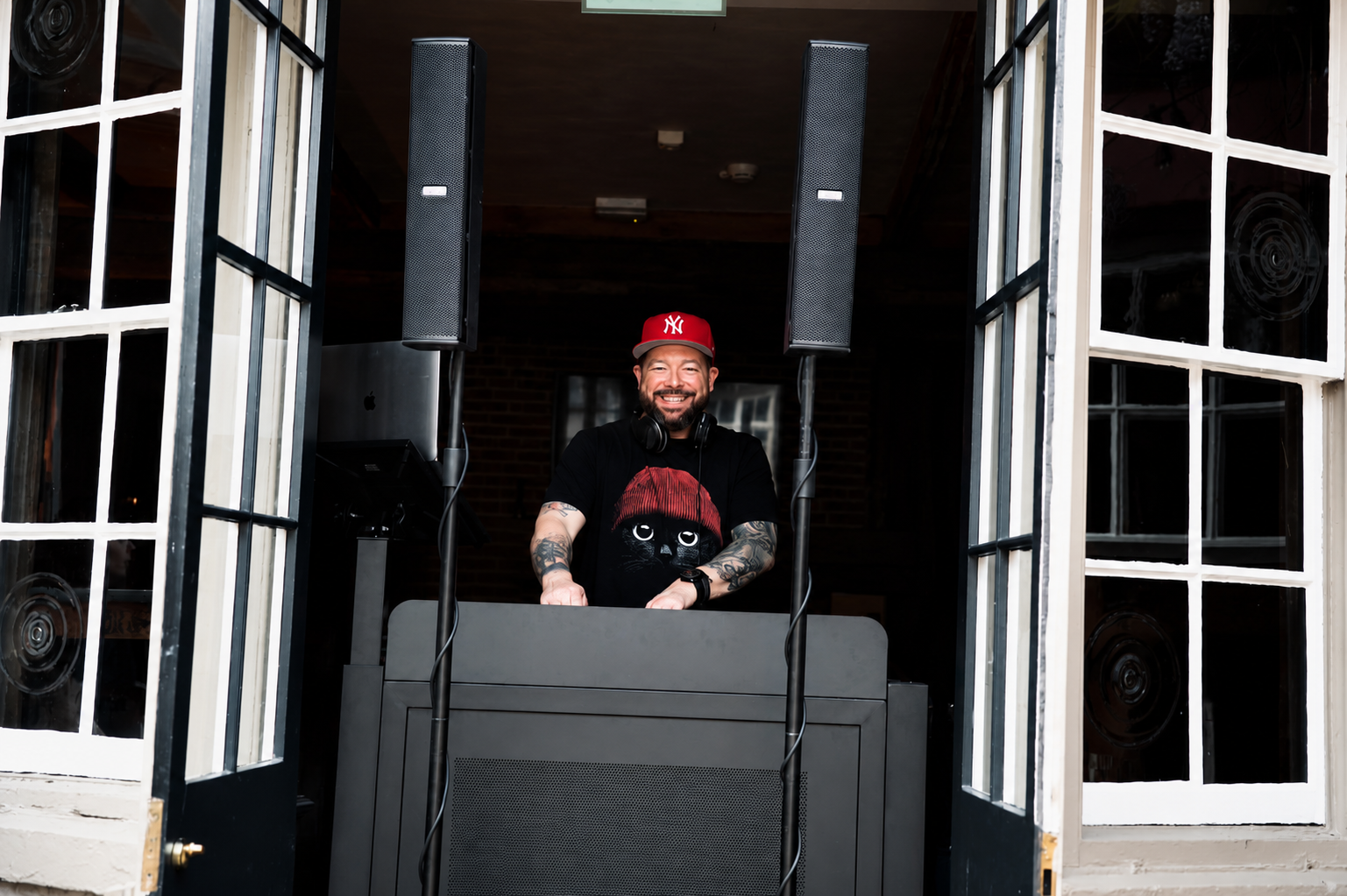 A smiling man with tattoos, wearing a red New York Yankees baseball cap and black t-shirt with a cat graphic, is DJing behind a booth with large speakers, inside a room with open window doors.