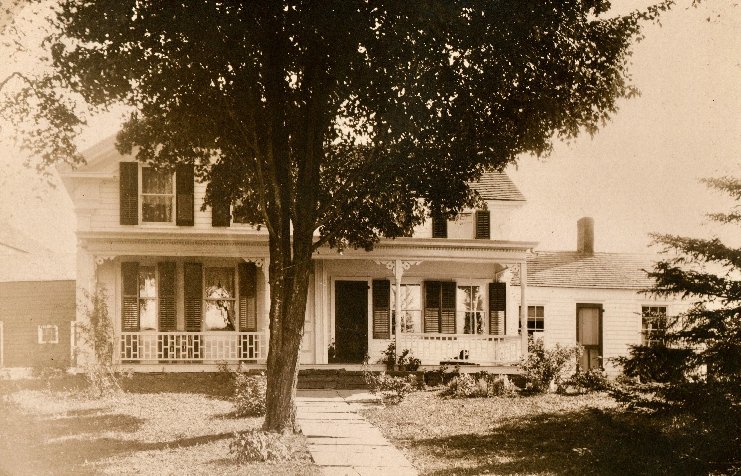 A vintage black-and-white photograph of a house with a large front porch, multiple windows with shutters, and a tree in front. The house has detailed trimwork and is surrounded by a well-kept lawn.