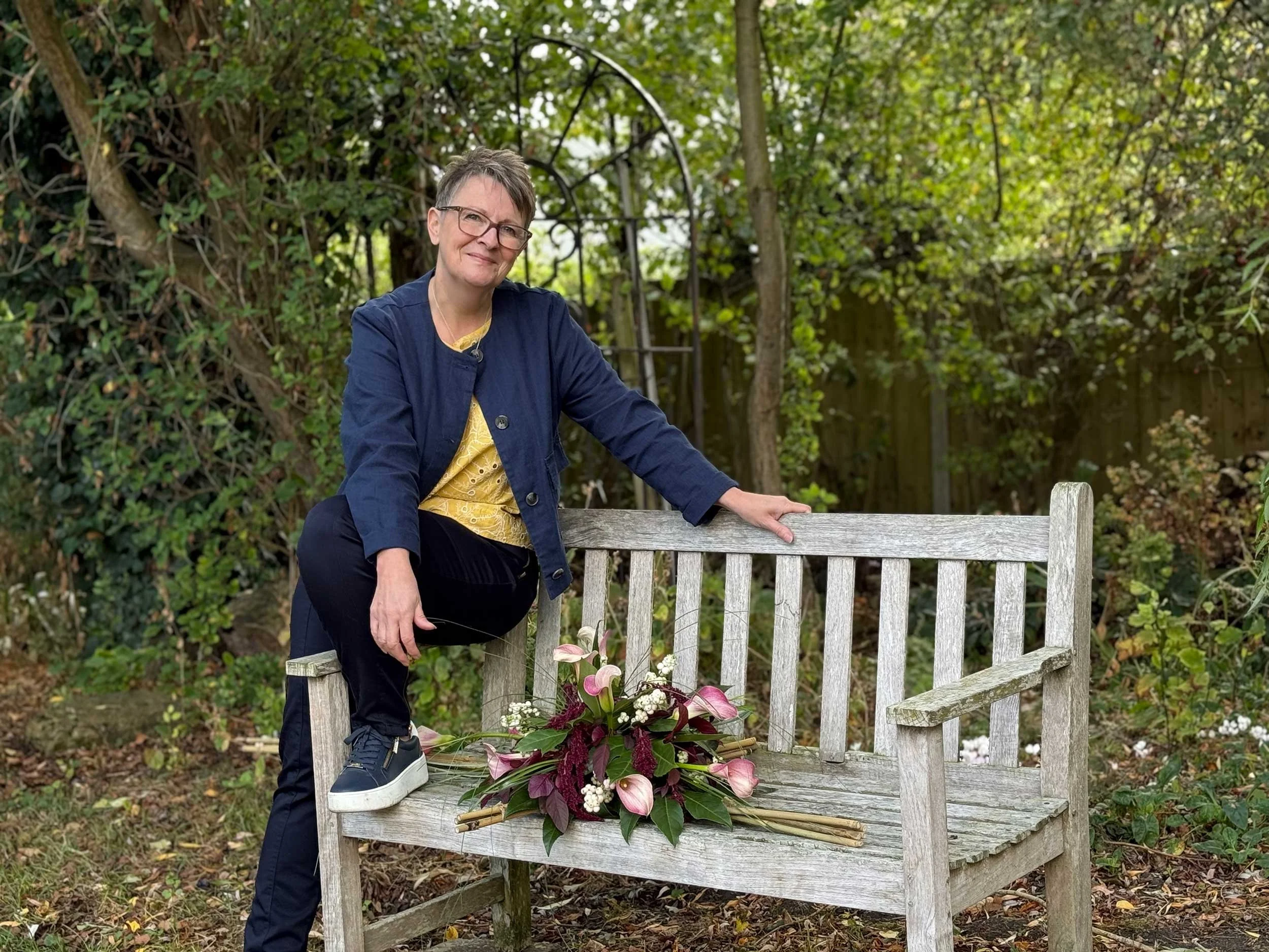 A woman with short gray hair and glasses sitting outdoors on a wooden bench, wearing a navy jacket and yellow shirt, with a bouquet of flowers in front of her, surrounded by green foliage.