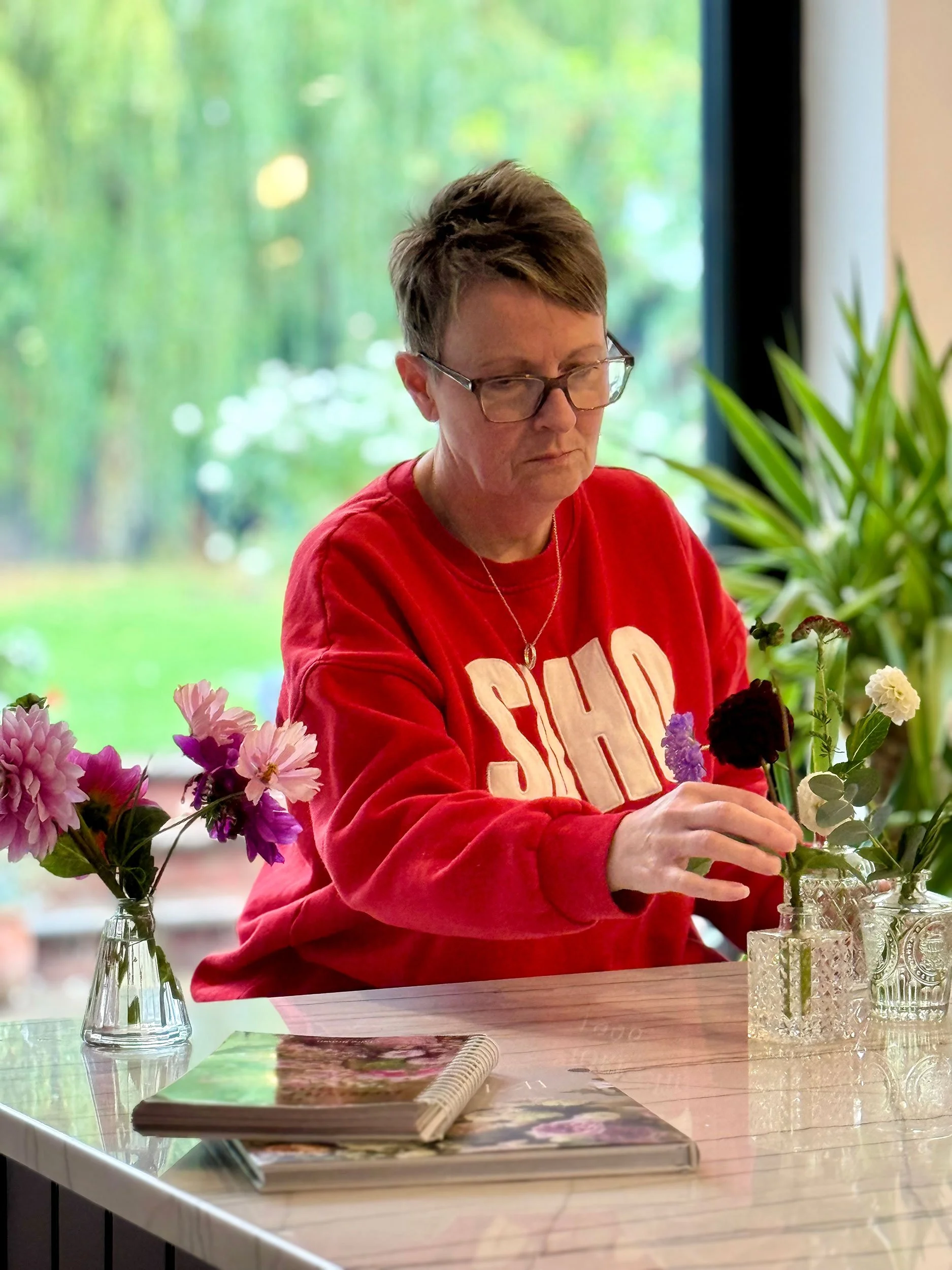 A woman with short brown hair and glasses, wearing a red sweatshirt, arranging flowers in a vase at a table with a window and green scenery in the background.