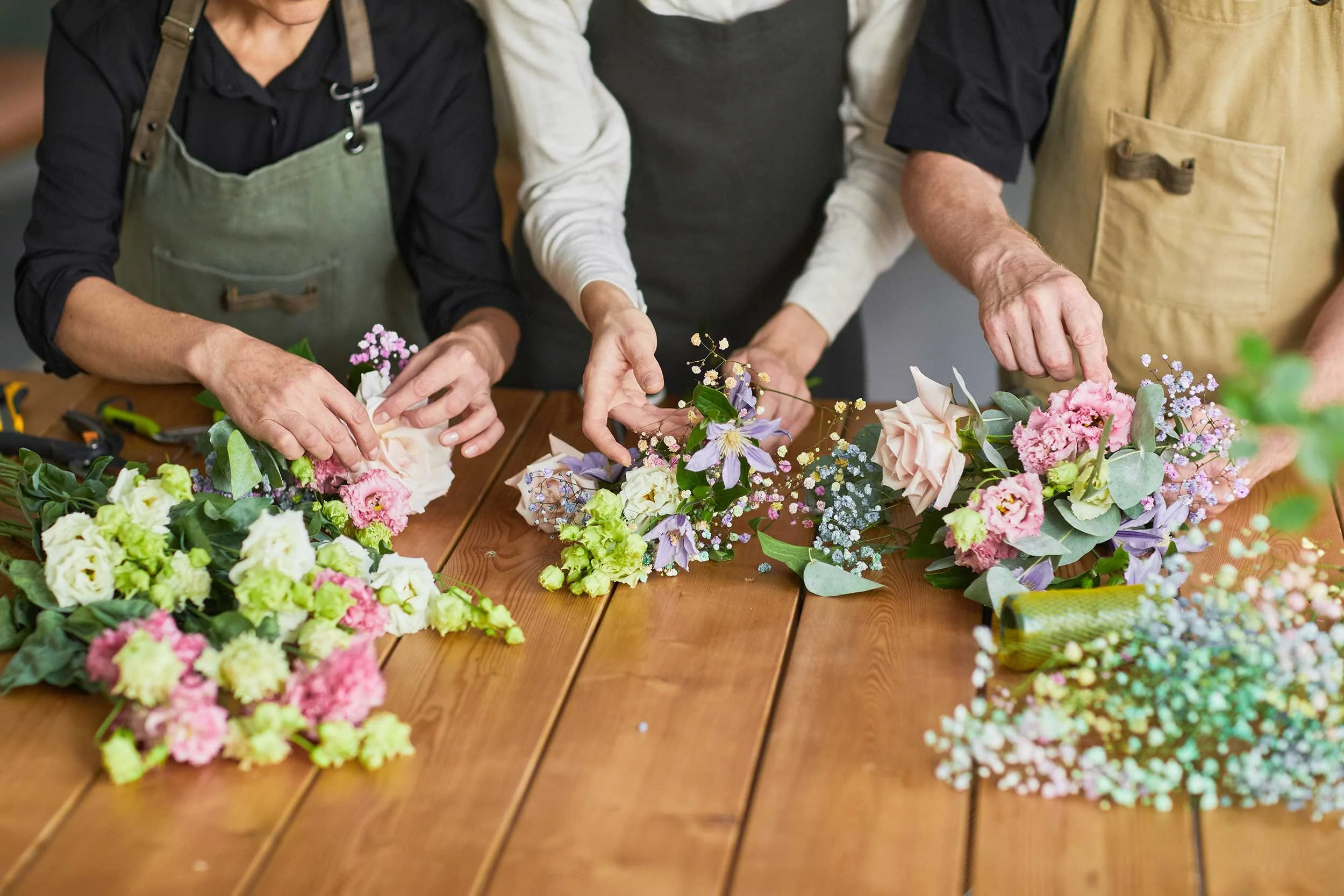 People arranging pink, purple, and white flowers on a wooden table.