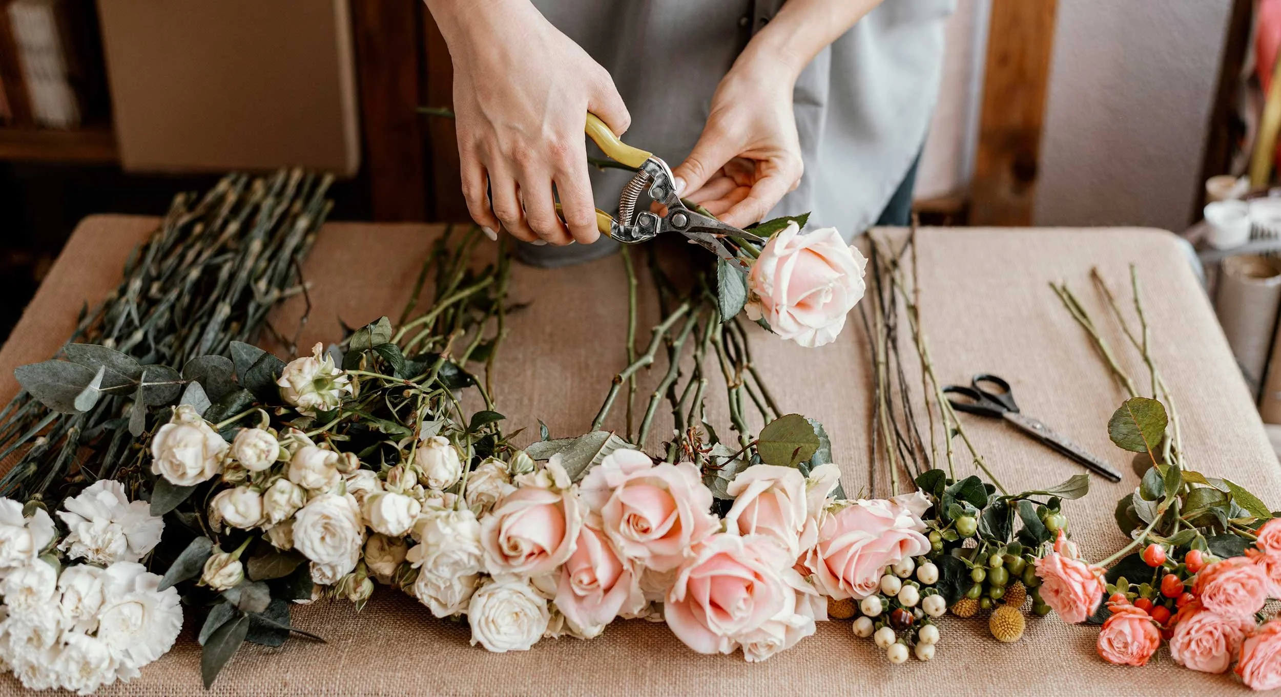 Person arranging pink and white roses on a table with floral supplies, including scissors and greenery.