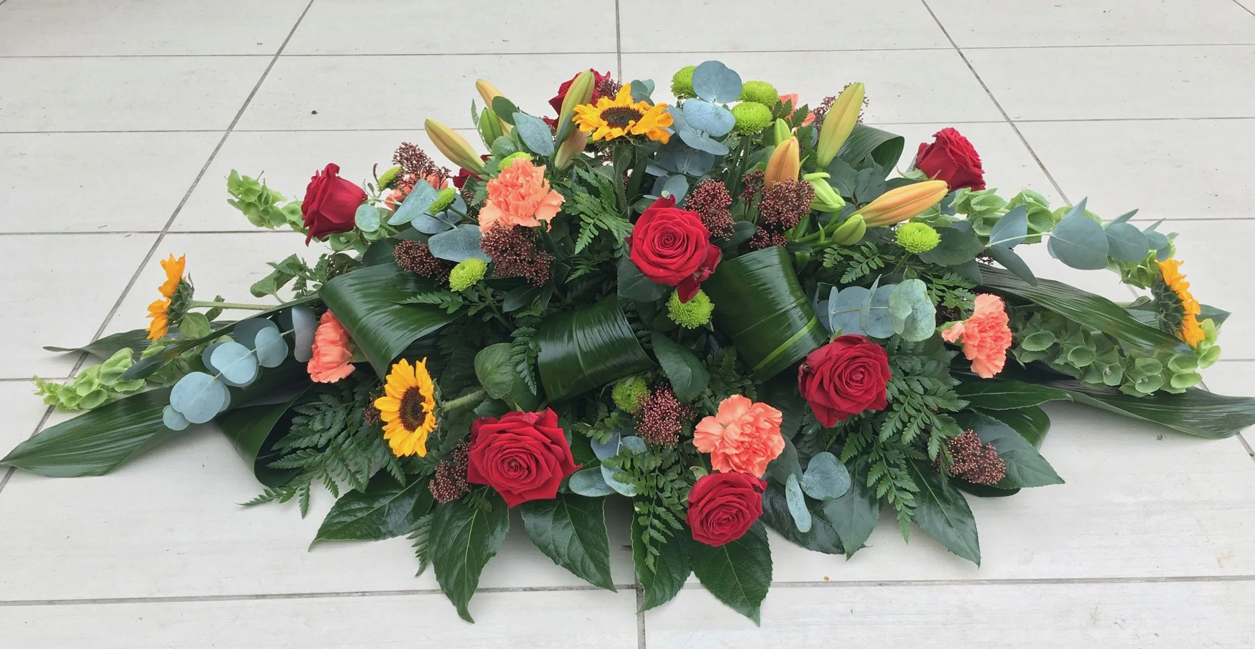 A floral arrangement with red roses, orange carnations, yellow lilies, sunflowers, green foliage, and eucalyptus leaves, laid on a tiled floor.