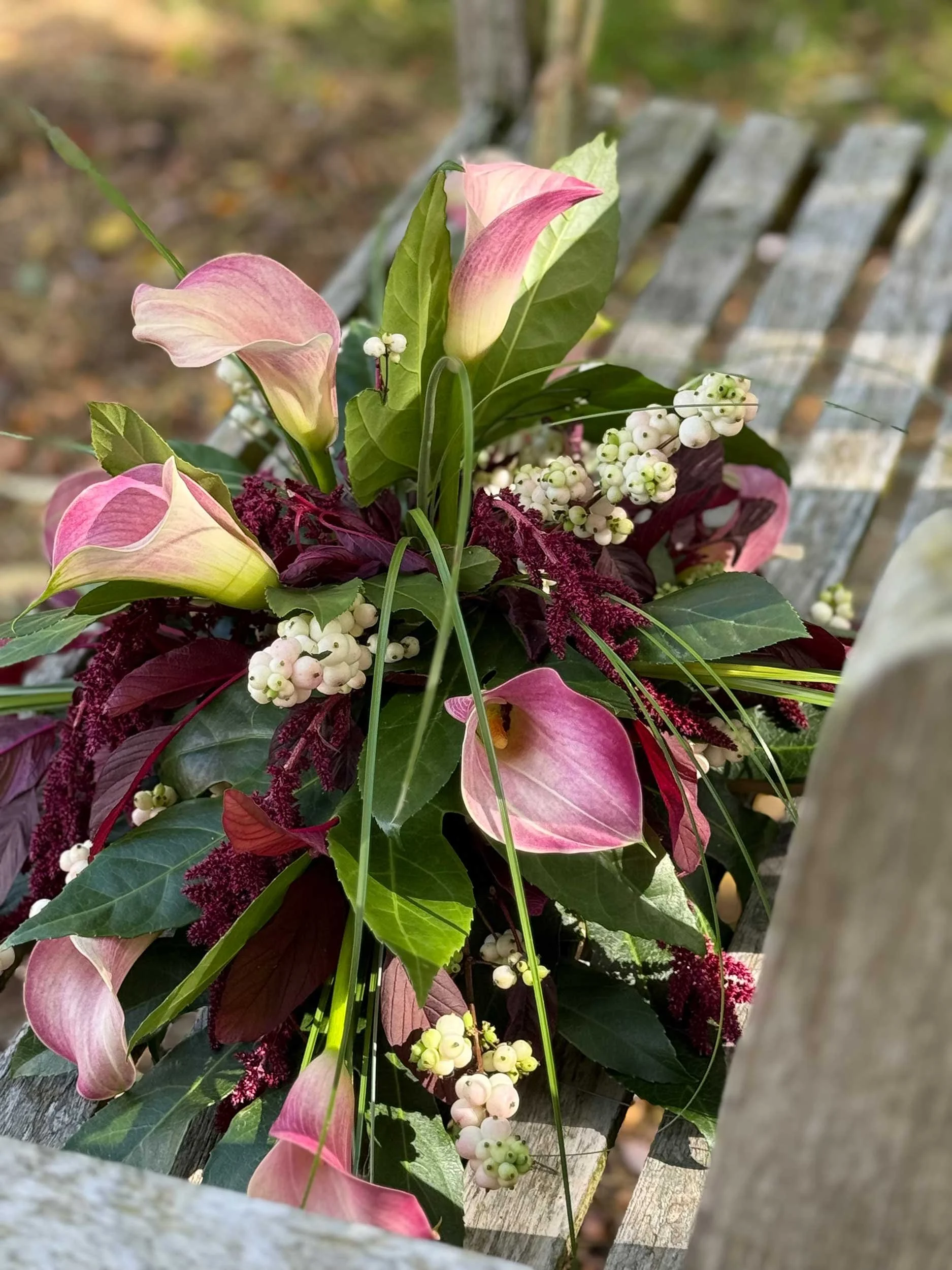 Close-up of a bouquet of pink and white calla lilies, white berries, and greenery placed on a wooden bench outdoors.