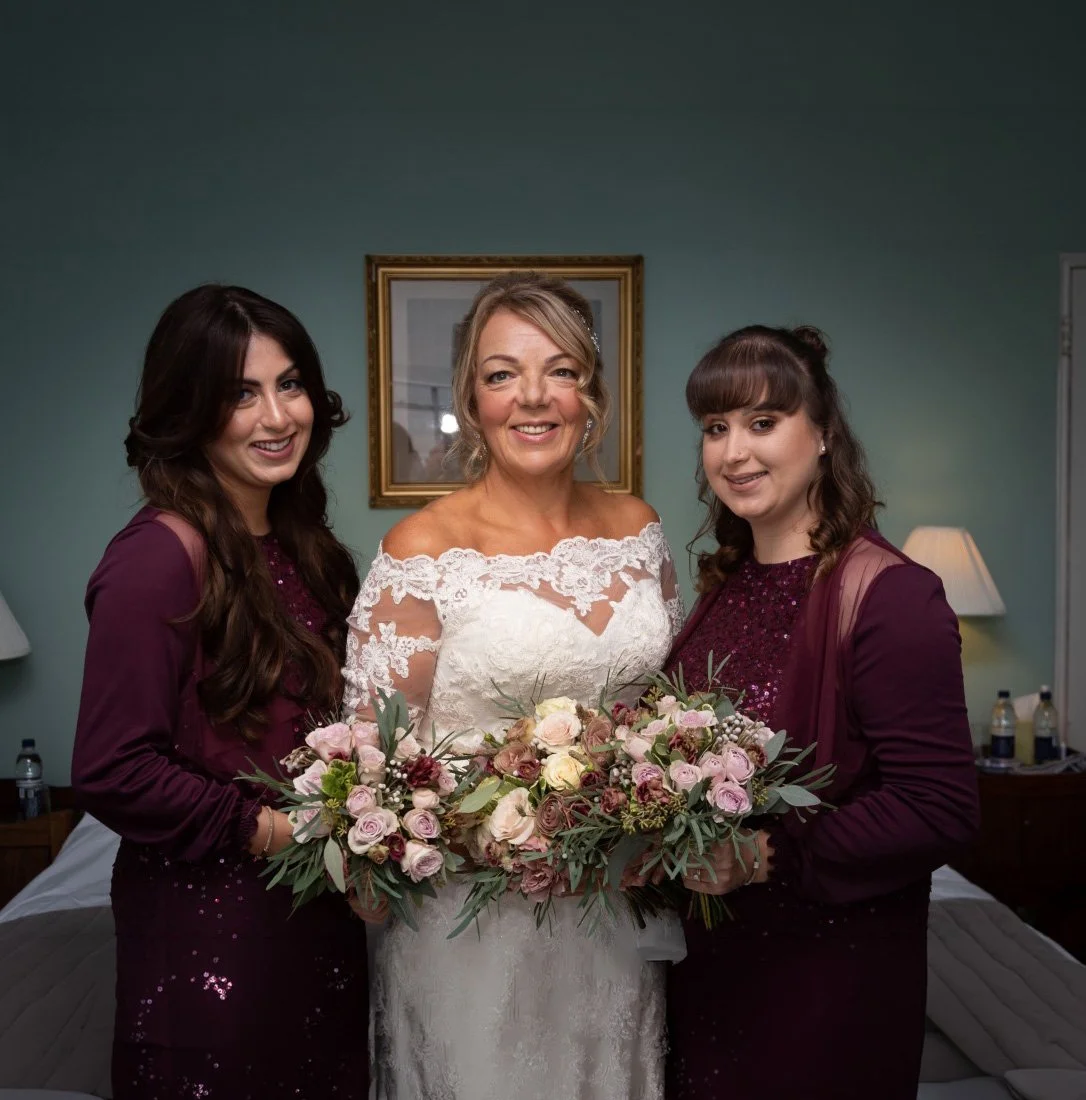 Three women, including a bride in a white lace wedding dress, smiling and holding bouquets of pink and white roses with greenery.