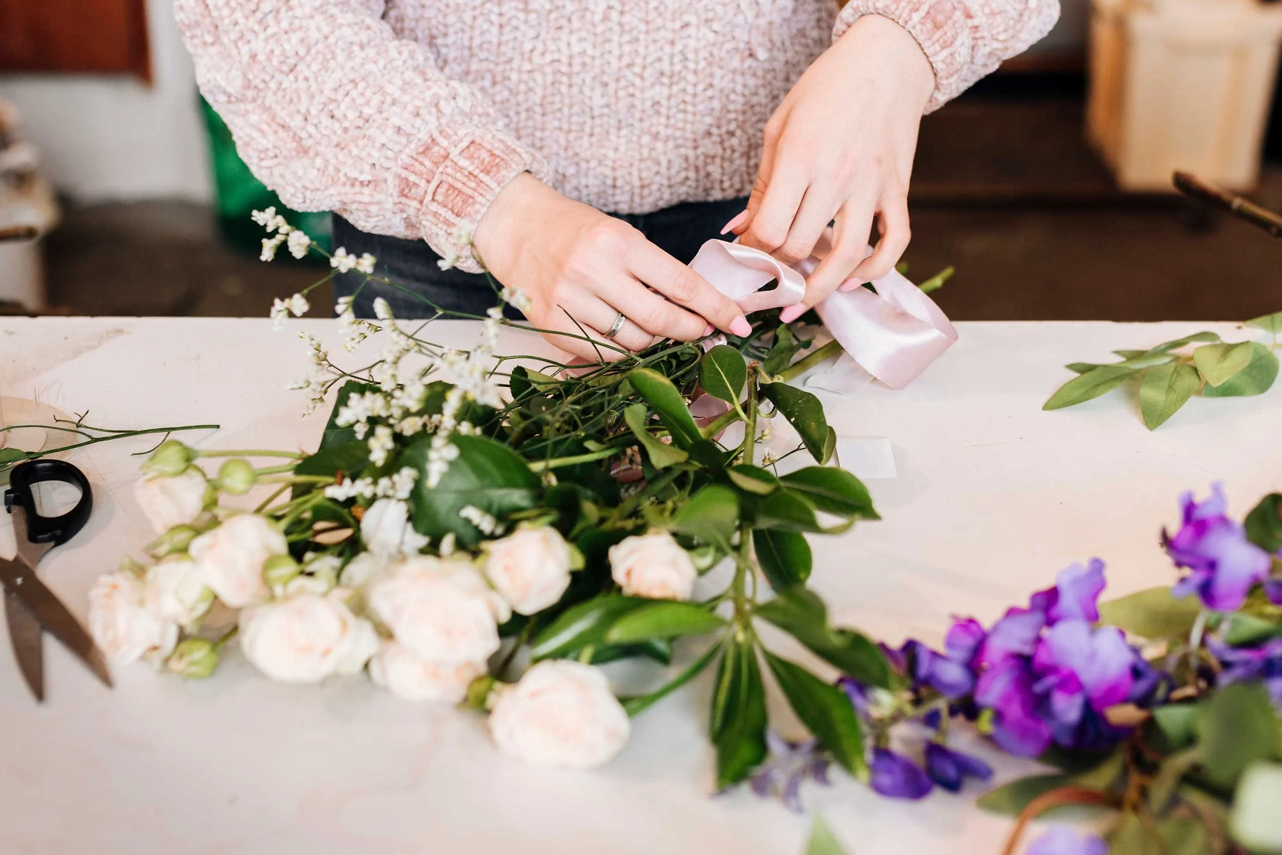 Person arranging pink satin ribbon on a bouquet of assorted flowers, including white roses, purple flowers, and greenery, on a white table.