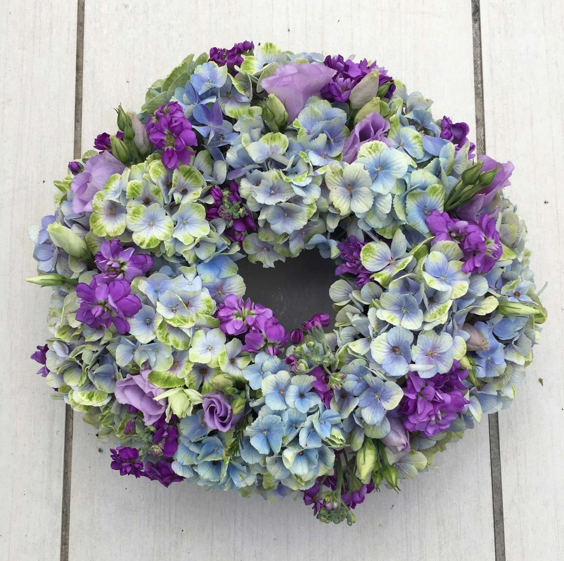 A heart-shaped floral arrangement with purple, blue, white, and green hydrangeas on a light-colored wooden surface.