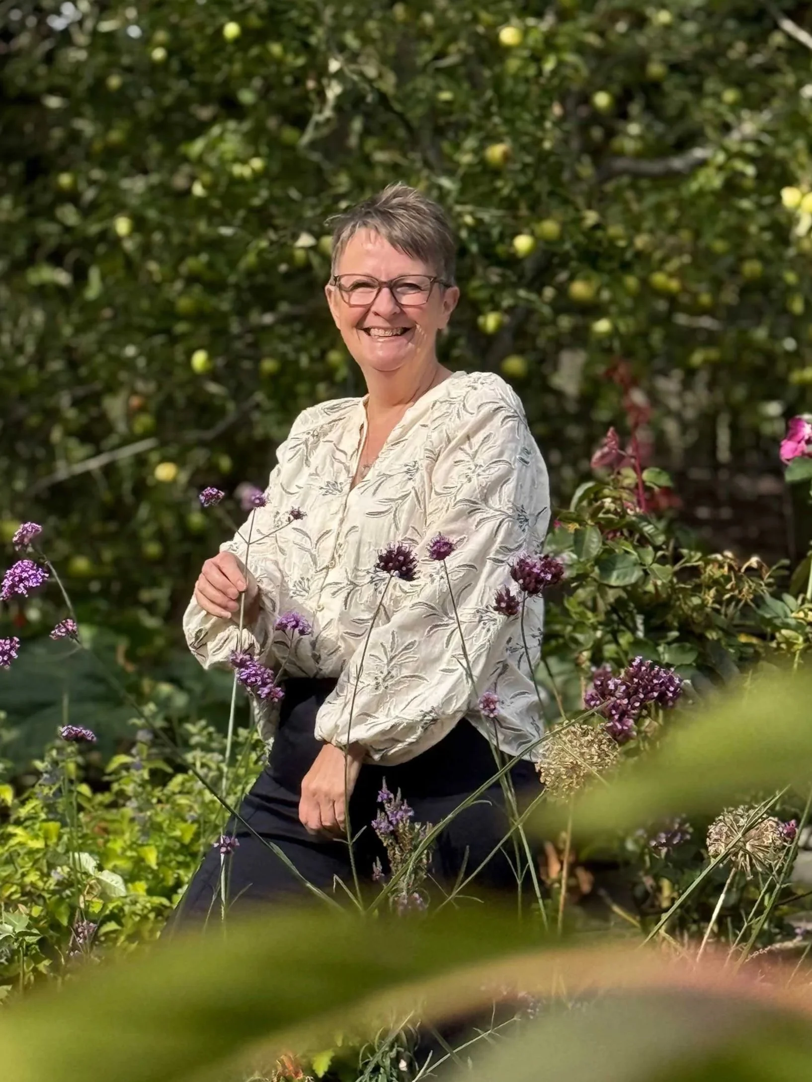 Smiling woman with short hair and glasses standing outdoors among flowering plants and greenery.