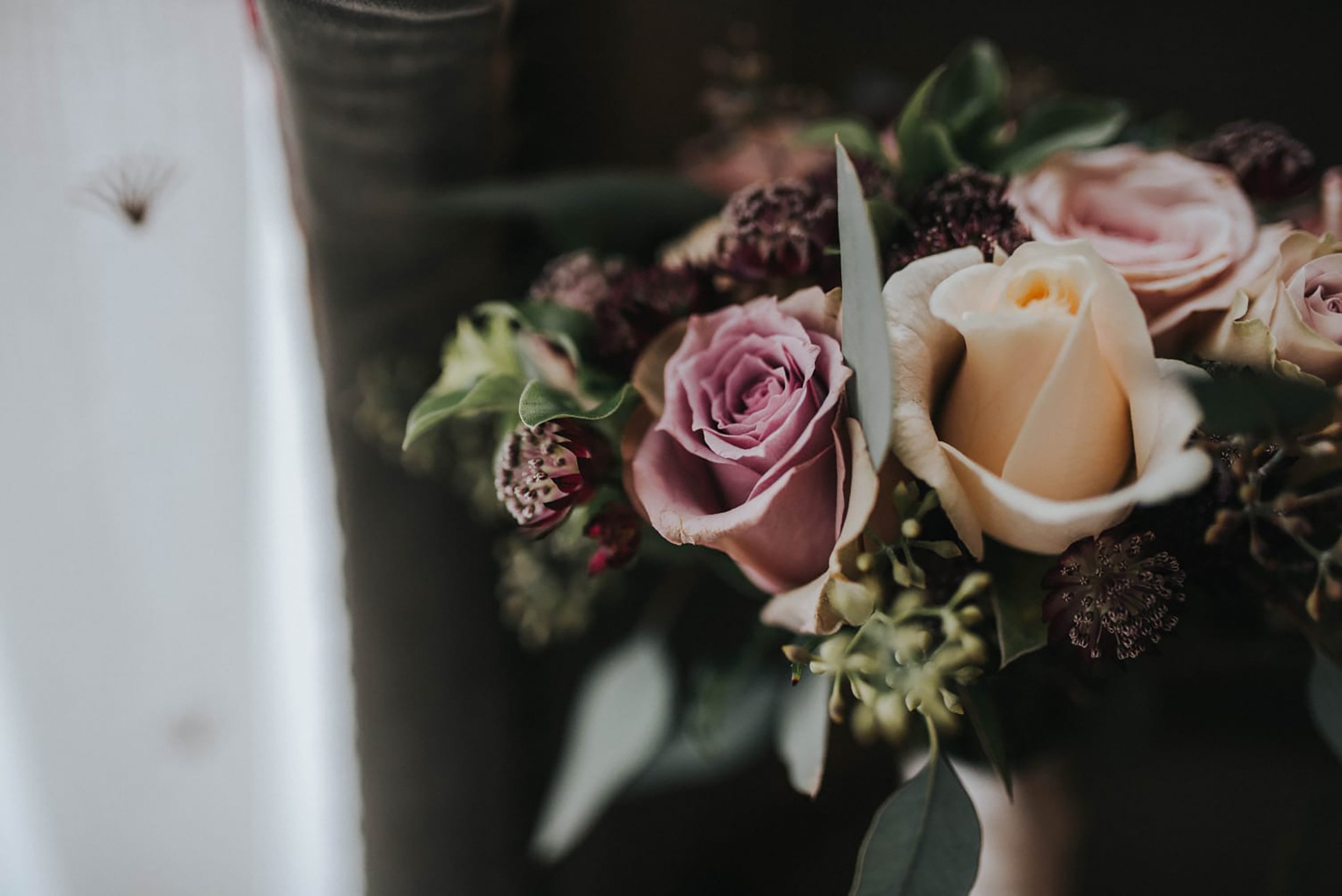 The image shows a close-up of a floral arrangement with pink, cream, and dark purple flowers, along with green leaves. It appears on a dark surface with a white background on the left side.