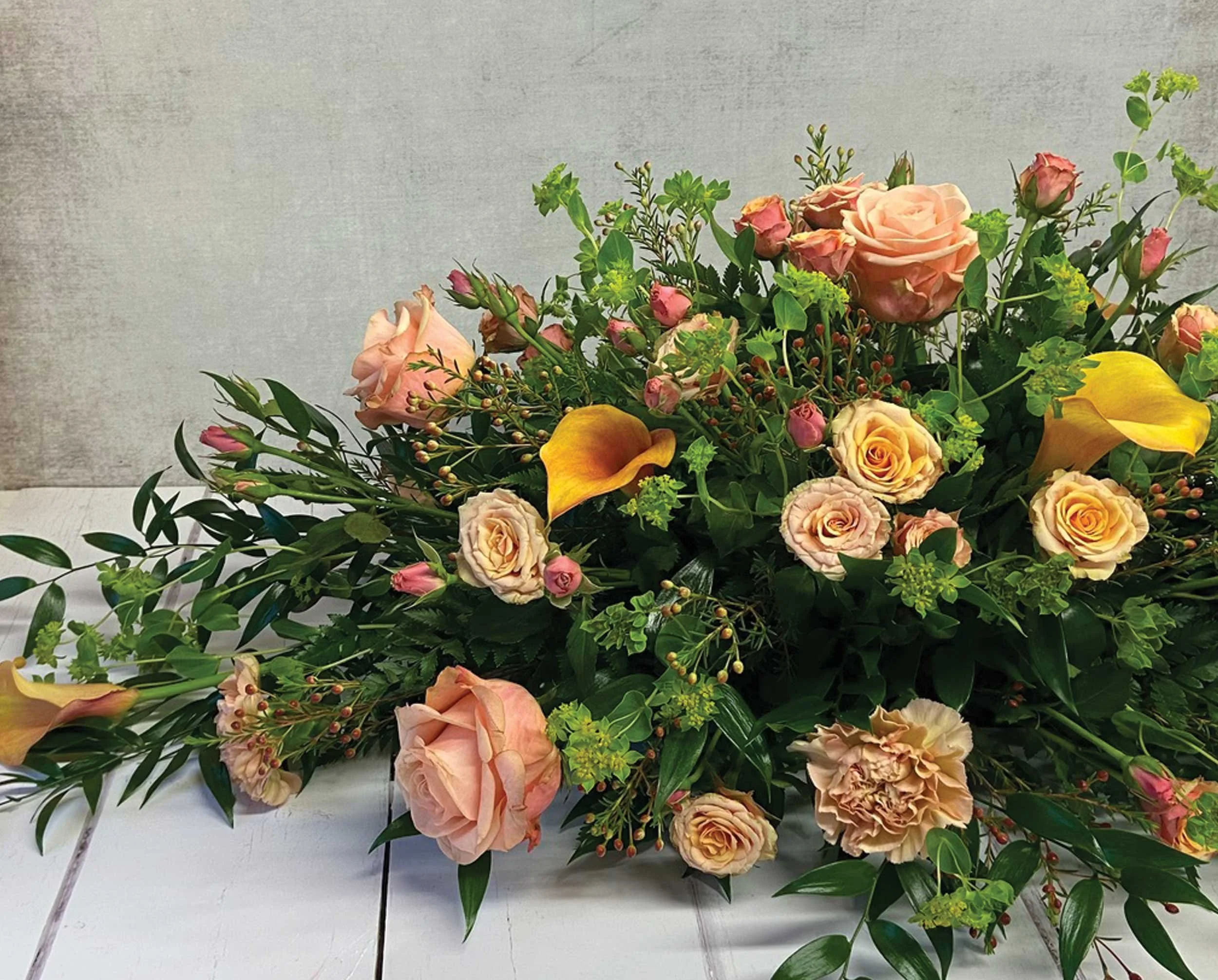 A floral arrangement featuring pink roses, yellow calla lilies, and assorted green foliage against a plain gray textured background.
