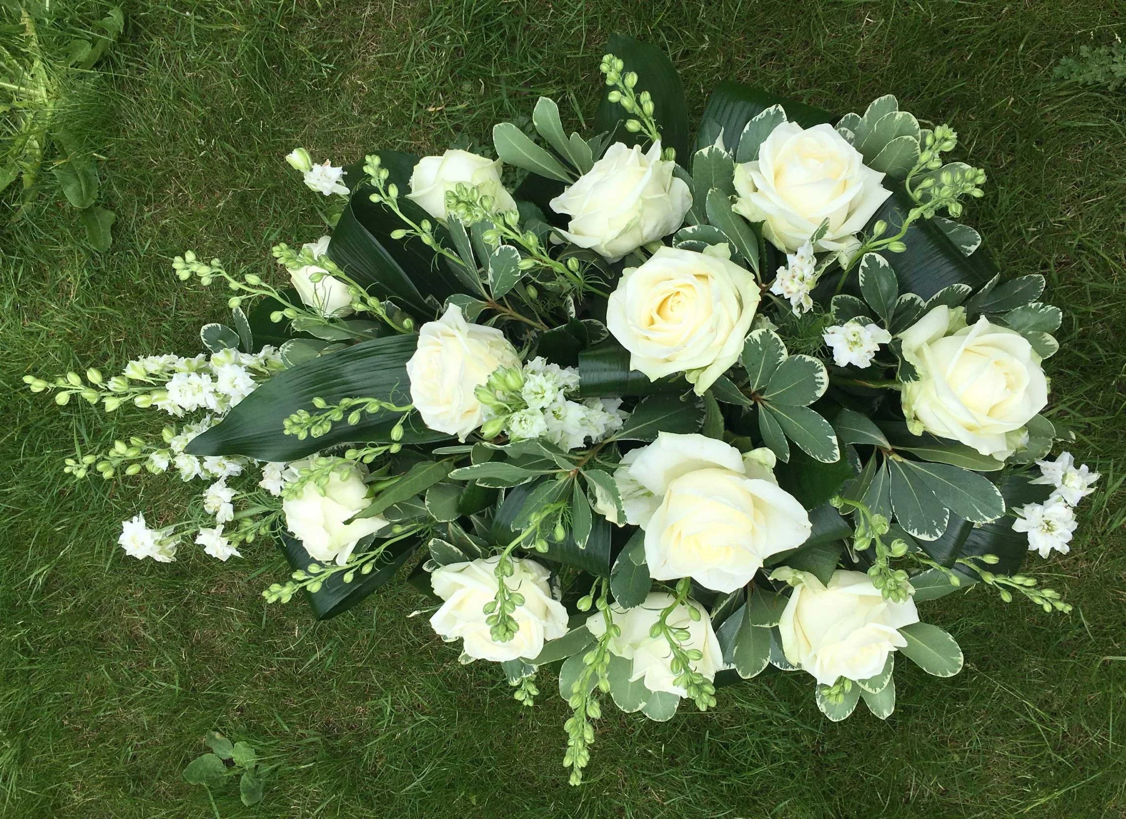 A floral arrangement of white roses, white stock flowers, and variegated leaves resting on grass.