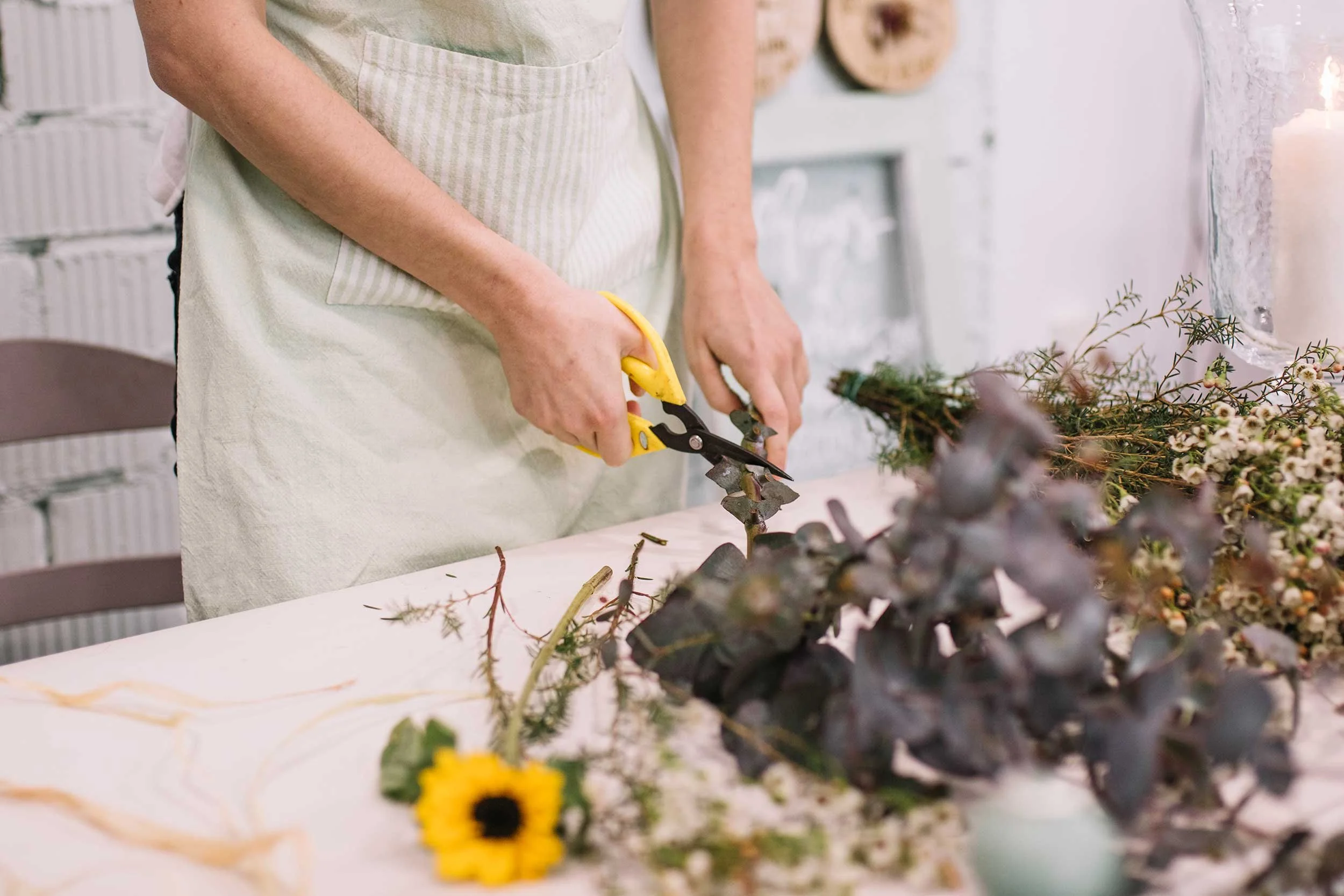 Person arranging flowers at a table, with sunflower, greenery, and other floral arrangements, in a room with white brick wall and candles.