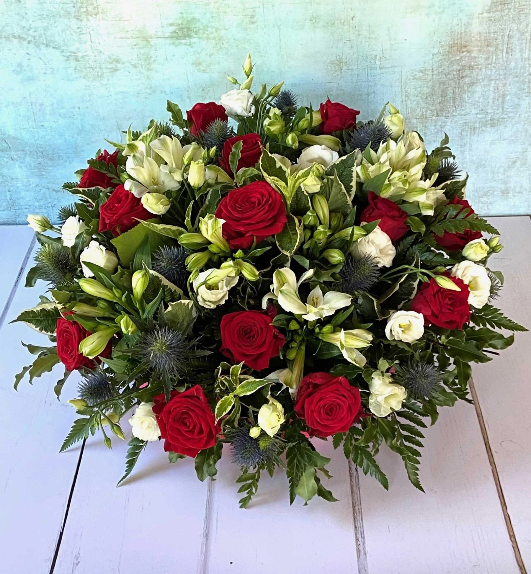 A bouquet of red roses, white lilies, white spray roses, and blue thistles arranged with green foliage on a white surface with a textured light blue background.