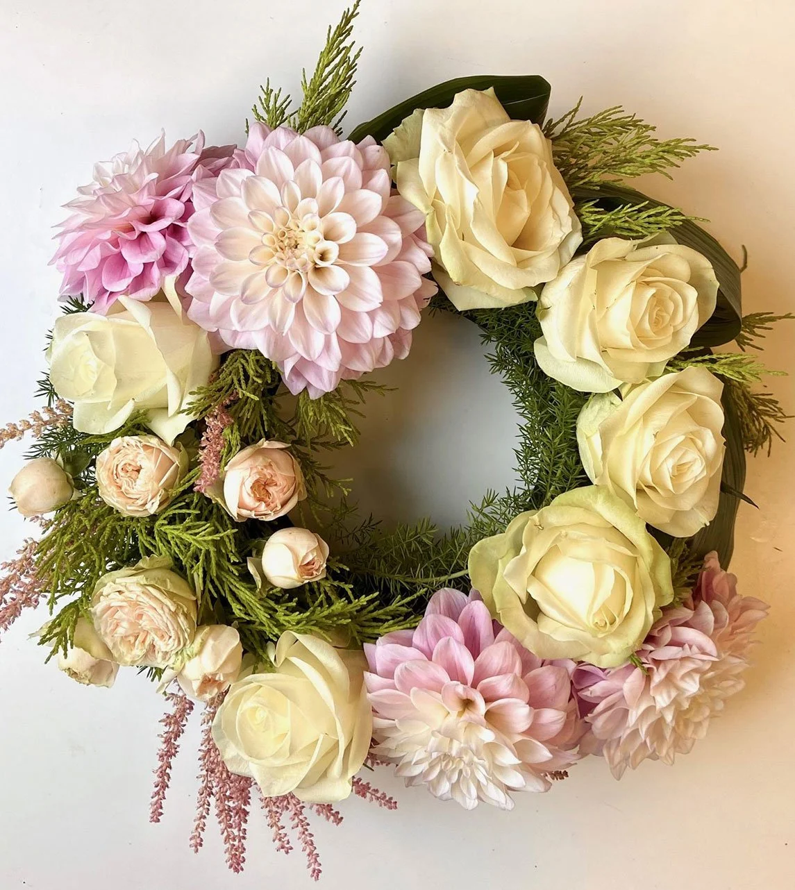 A floral wreath consisting of white roses, pink dahlias, smaller pink flowers, and greenery, arranged in a circular pattern.