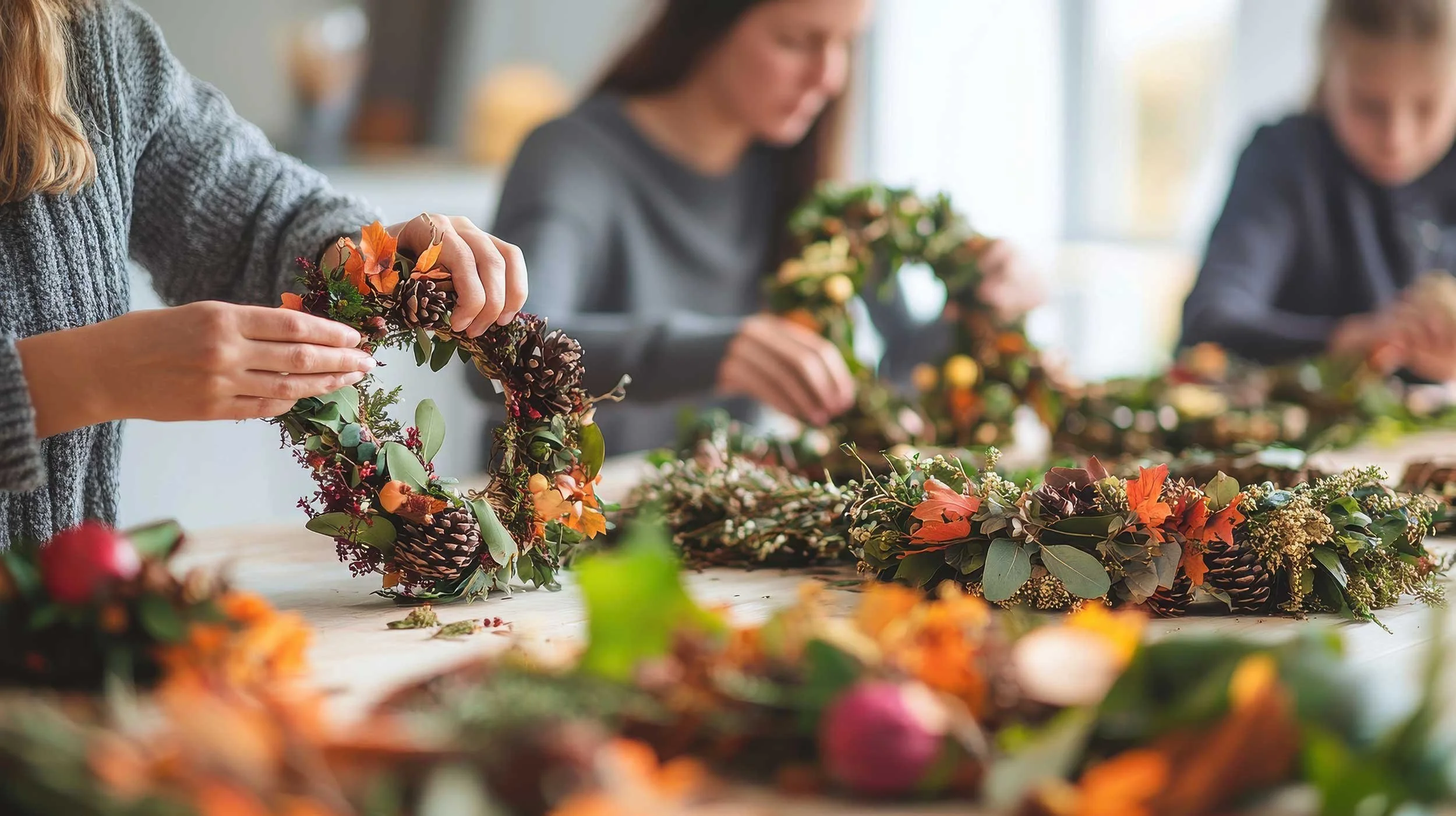 People working on making autumn-themed floral wreaths with pinecones, leaves, and flowers at a table.