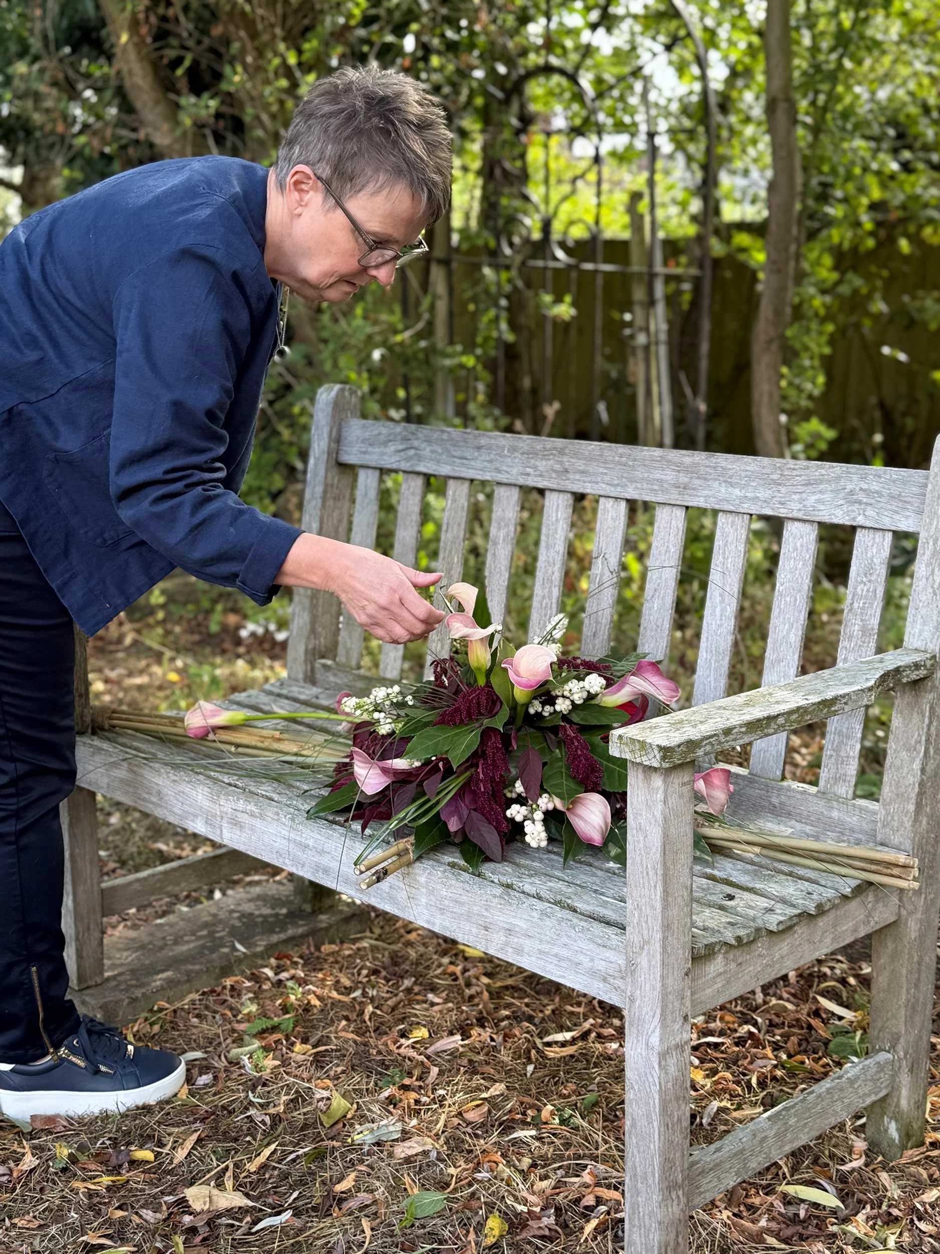 A person with short brown hair, glasses, wearing a blue jacket, leans over and arranges flowers on a weathered wooden garden bench outdoors. The bench has a bouquet of pink and white flowers, including white berries and dark green and burgundy leaves. The background includes trees, a wooden fence, and fallen leaves on the ground.