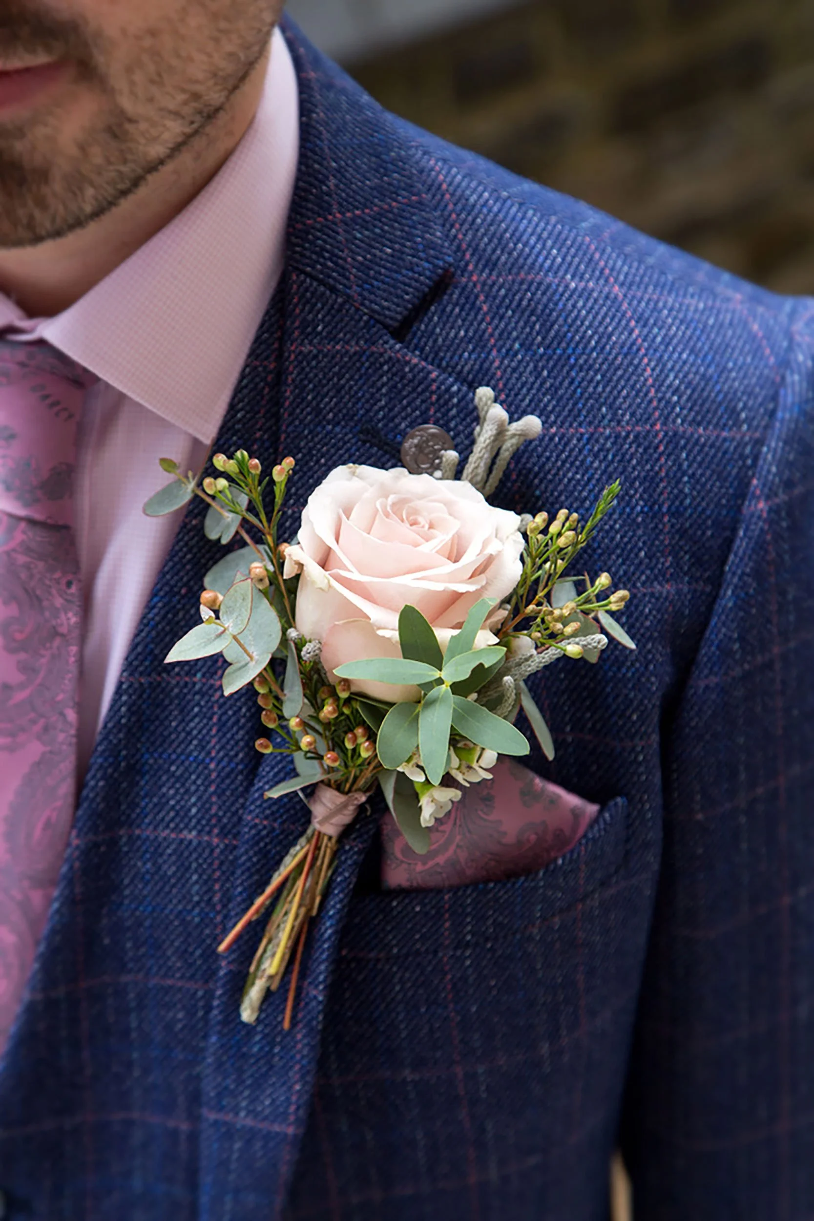 Close-up of a man's suit with a pink shirt and a boutonnière featuring a light pink rose and greenery.