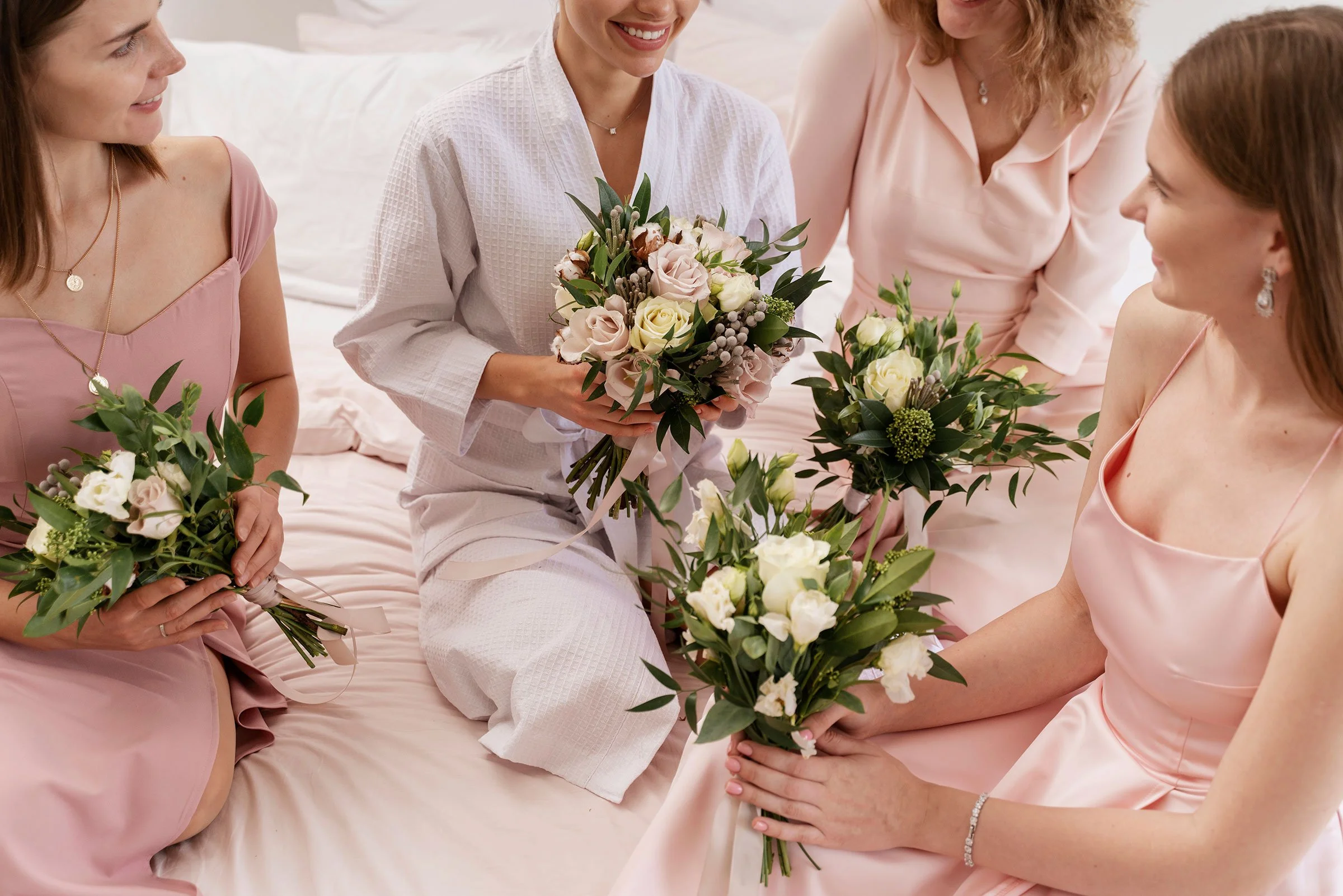 Four women sitting on a bed, dressed in pink and white attire, holding bouquets of flowers, smiling, and participating in a celebration or gathering.