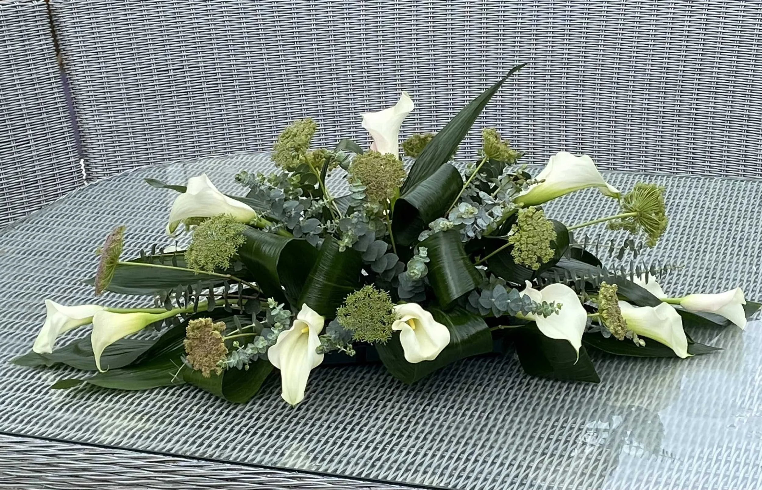 A floral arrangement with white calla lilies, green hydrangeas, eucalyptus leaves, and long green leaves, placed on a glass table with a wicker background.