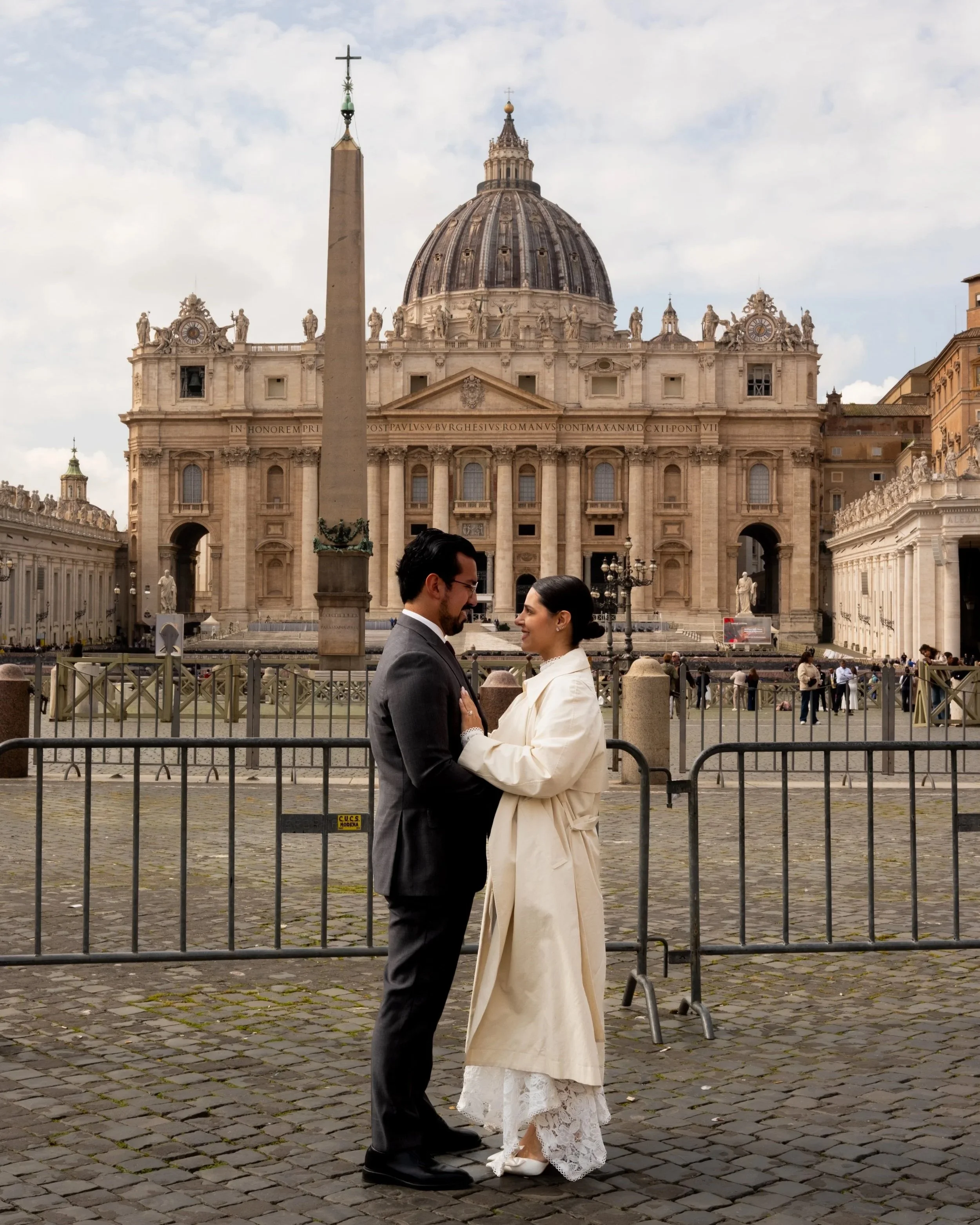 A couple in wedding attire standing close together and smiling, with St. Peter's Basilica in Vatican City in the background.