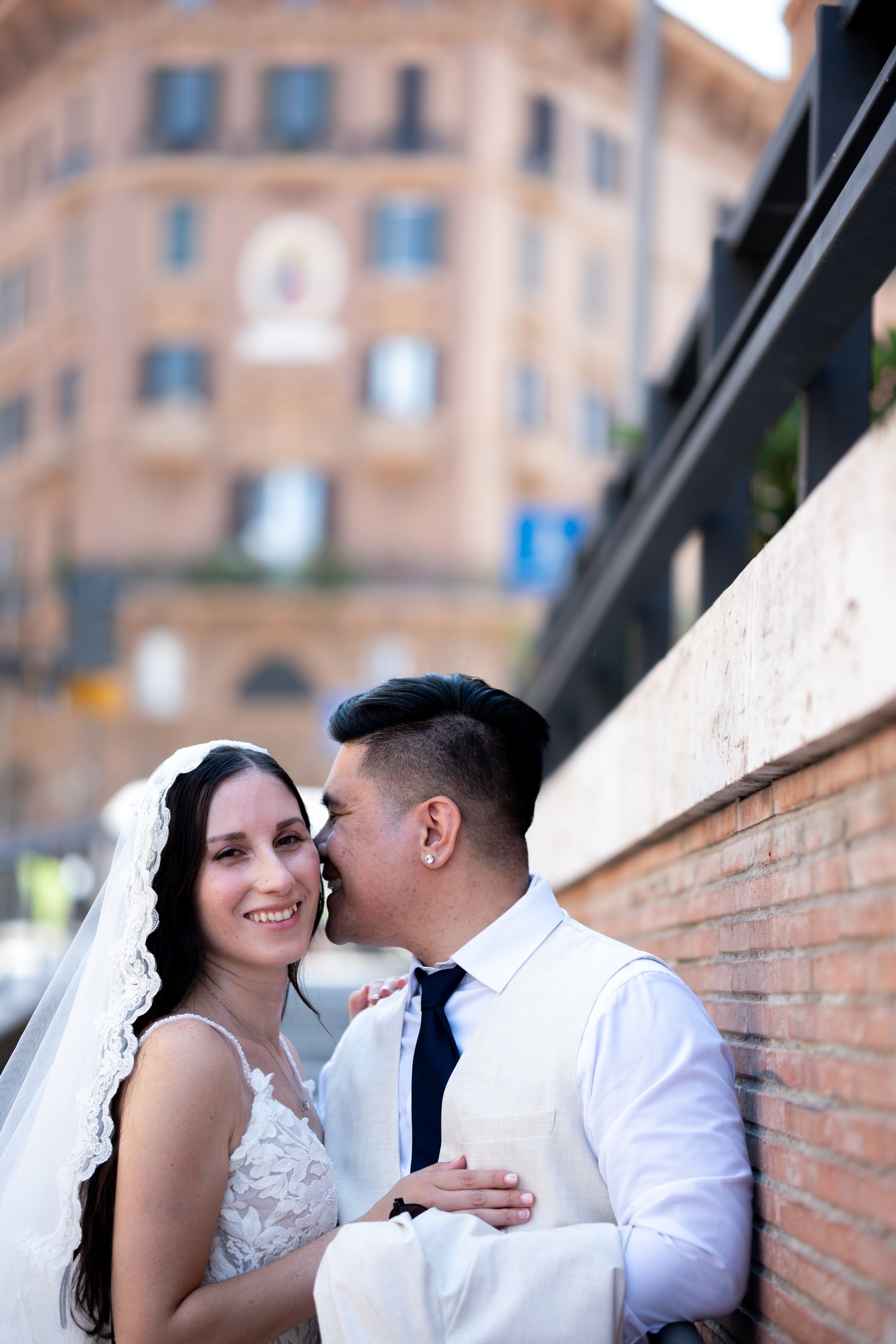 A bride and groom are close together, smiling, with the groom whispering in the bride's ear. The bride is wearing a white lace wedding dress and veil, while the groom is in a white suit with a navy tie. They are standing outdoors against a brick wall with a blurred background of a building