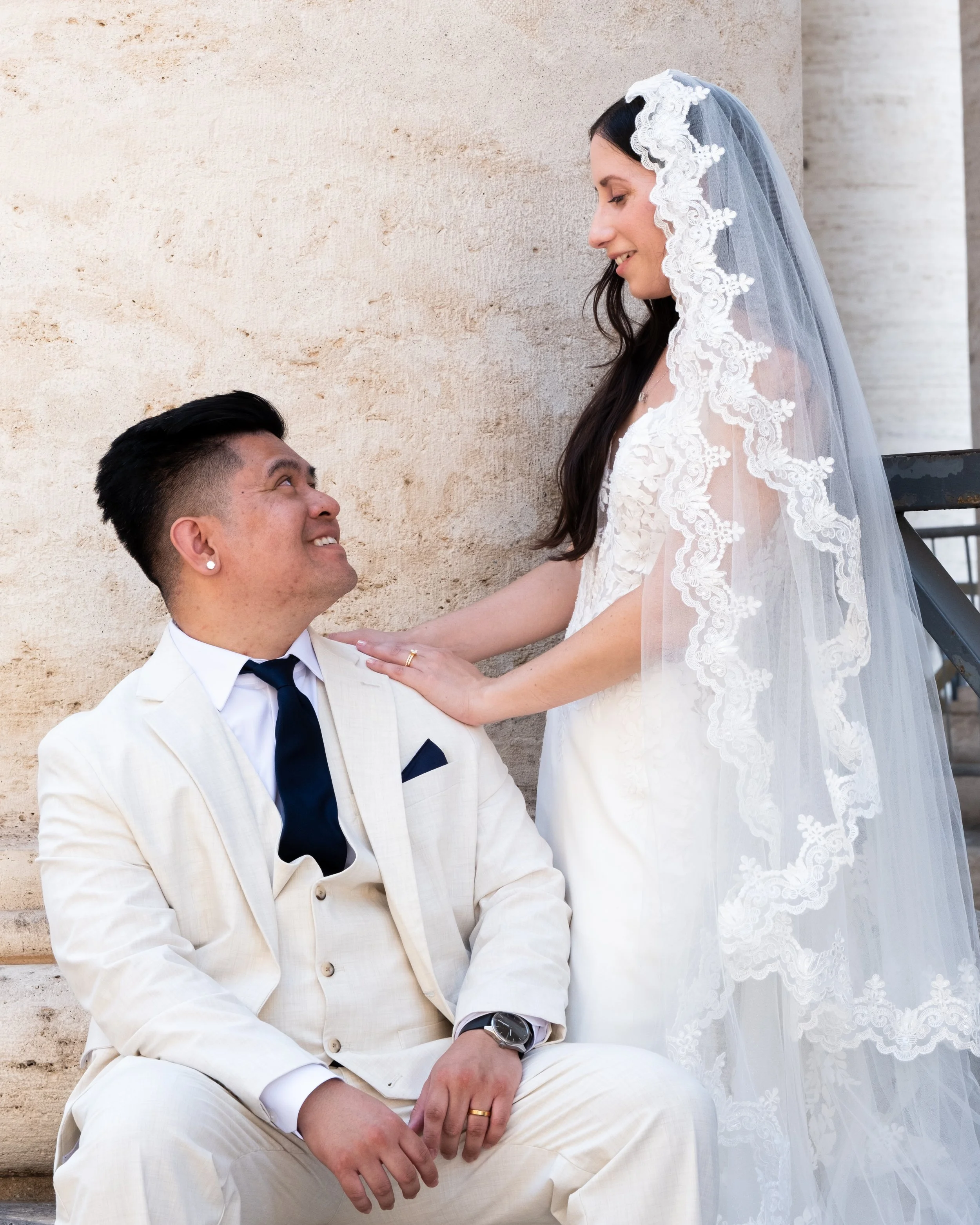 A man in a white suit and black bow tie sitting on stairs and looking up at a woman in a wedding dress and veil standing in front of him, smiling.