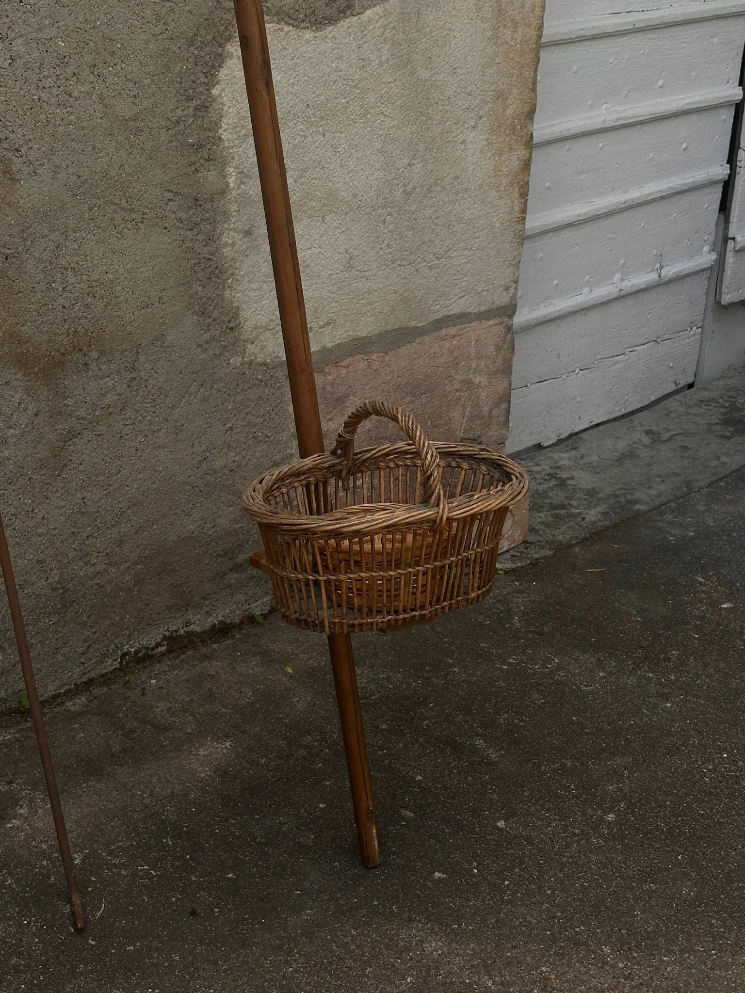 Panier en osier suspendu sur un support en bois avec un fond de mur en béton et une porte en bois blanc.