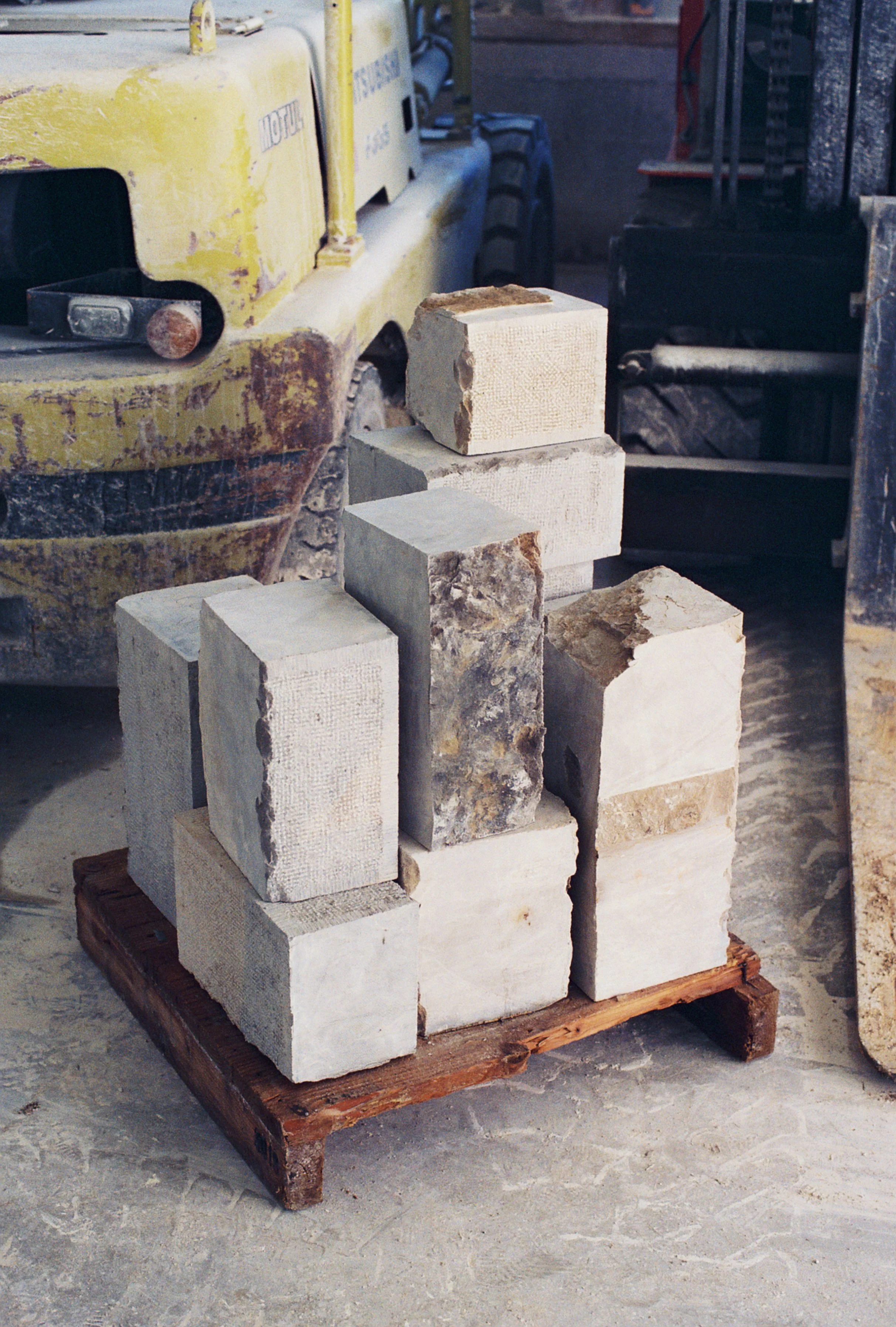 Un treillis de blocs de pierre empilés sur une palette en bois, avec un véhicule jaune à l'arrière et un sol en ciment ou pierre serapée.