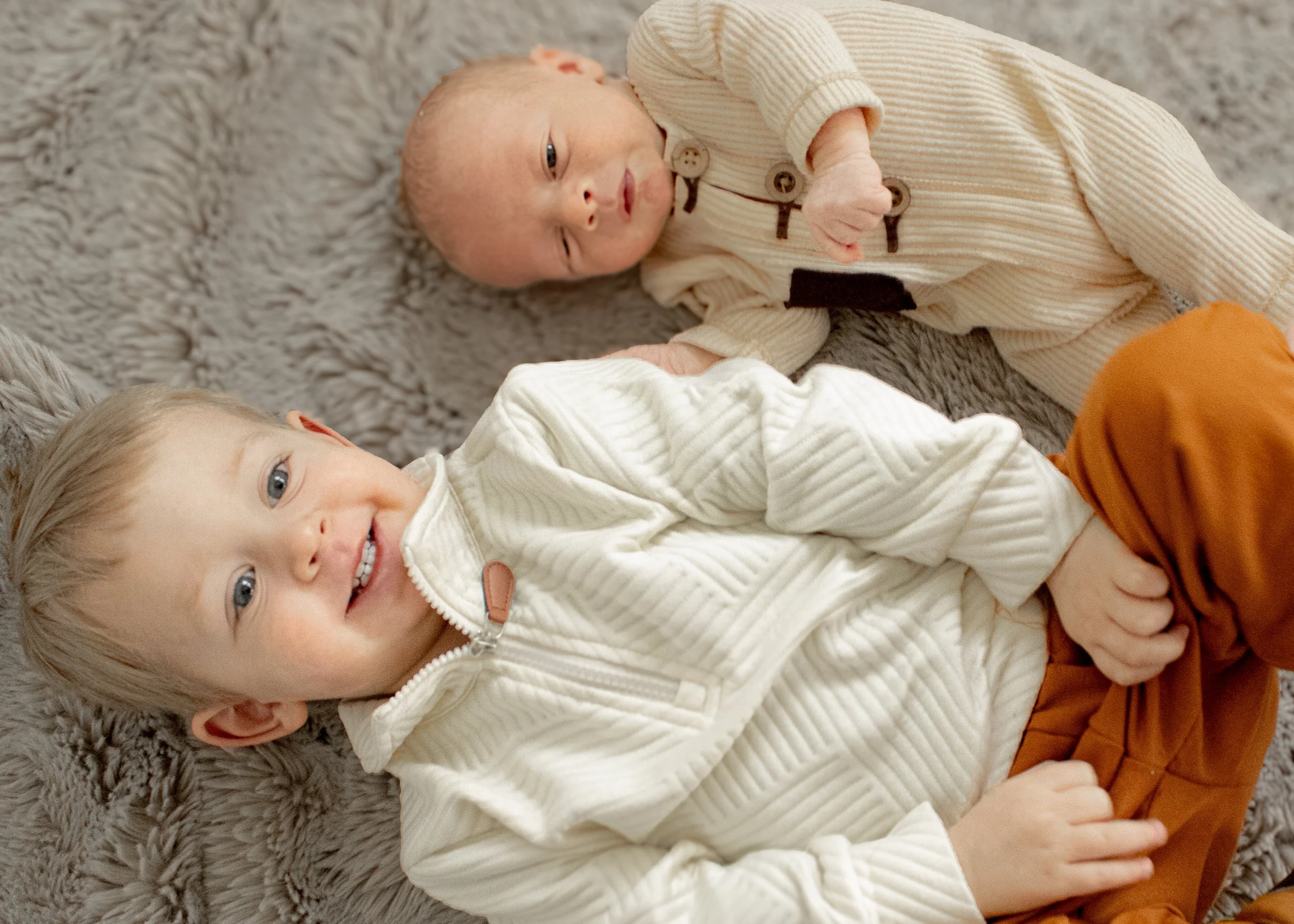 Two young children lying on a soft, gray, plush carpet. One child has blonde hair and is smiling at the camera, wearing a light-colored, zip-up jacket. The other child has a newborn-like appearance with a slightly closed eye, dressed in a beige, stri