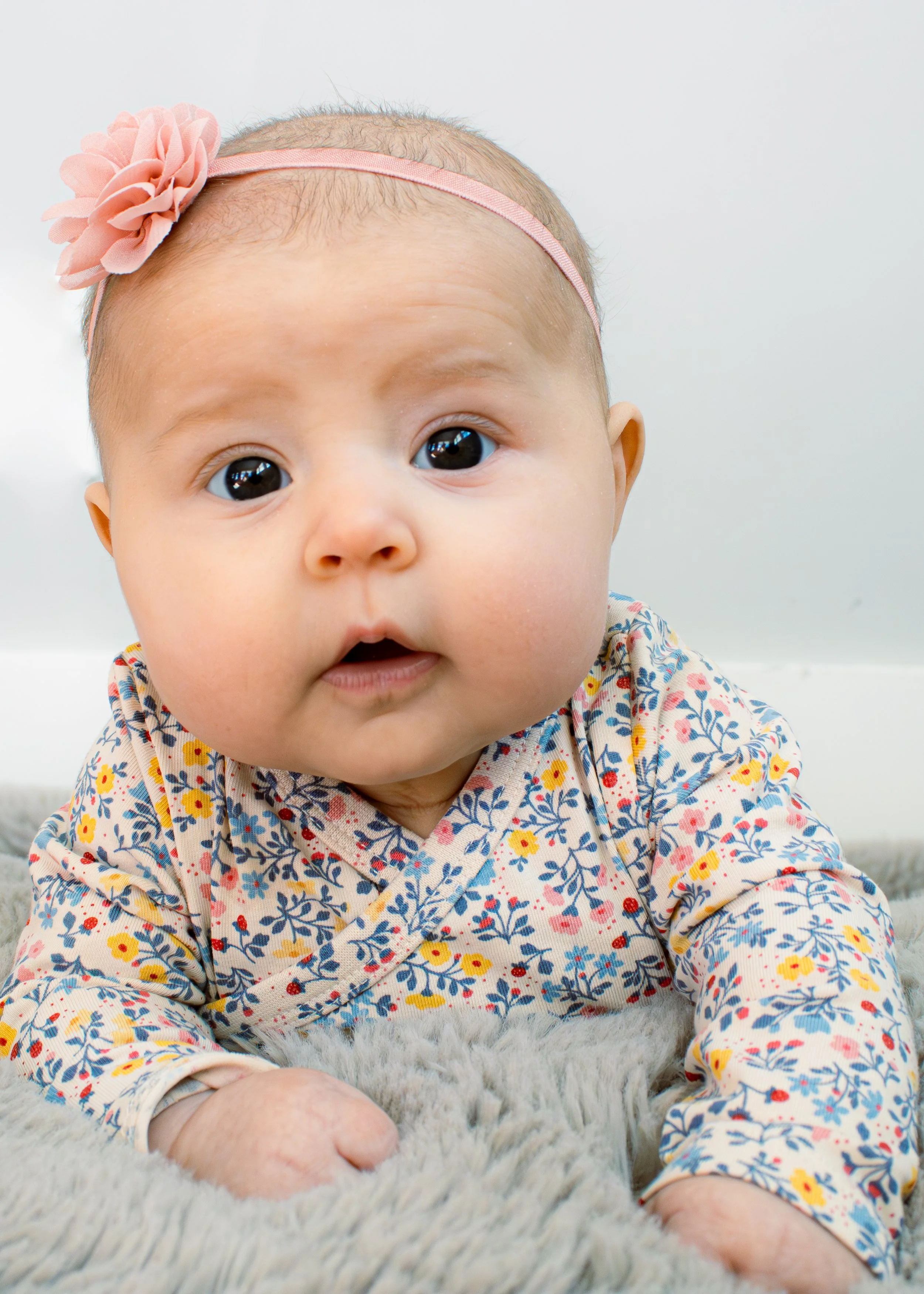 Close-up of a baby lying on a soft blanket, wearing a colorful floral dress and a pink headband with a flower, with large dark eyes looking at the camera.