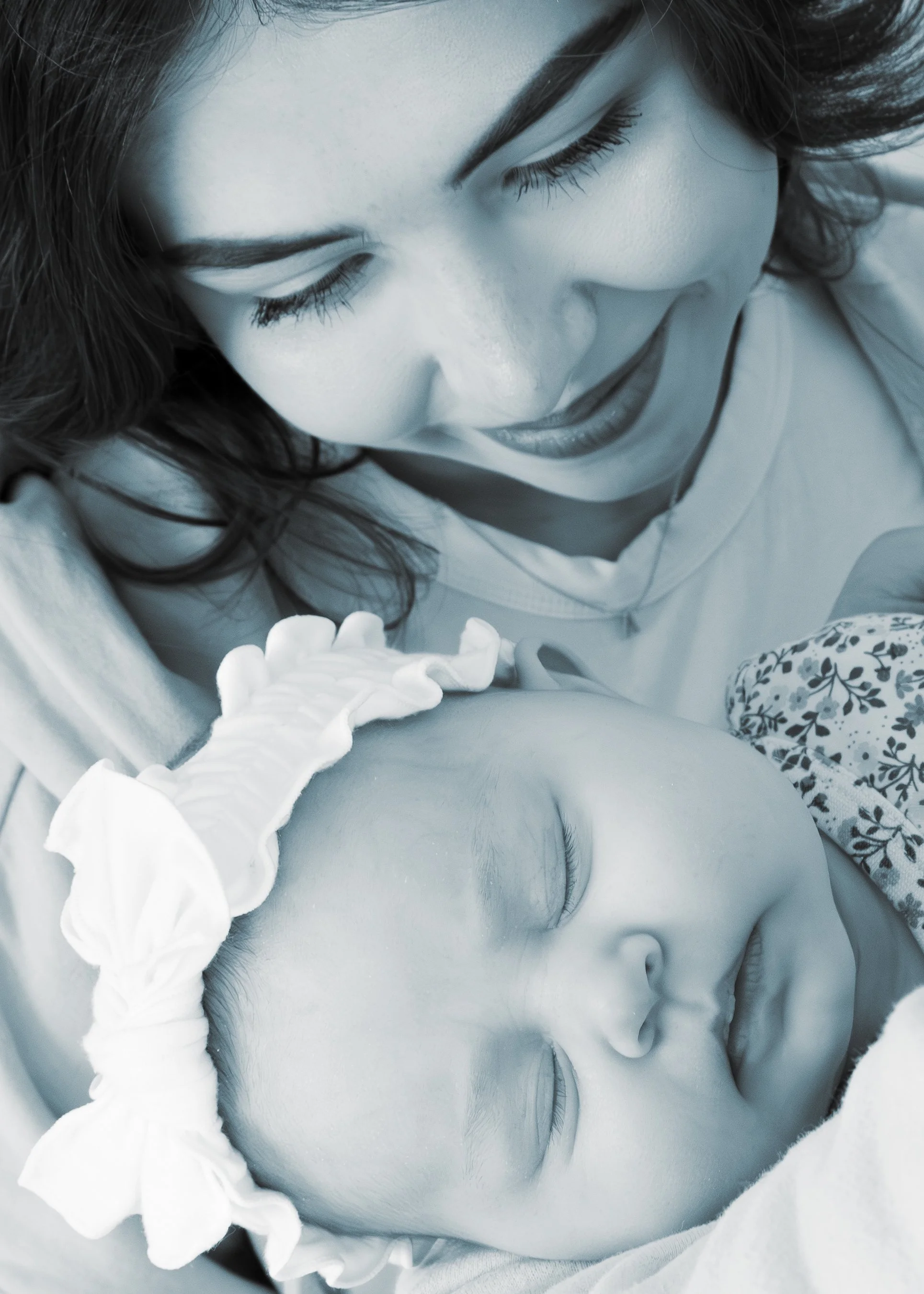 A woman with dark hair and closed eyes, wearing makeup, smiling with a baby lying on her, wearing a light-colored top and ruffled sleeves, in a close-up black and white photograph.