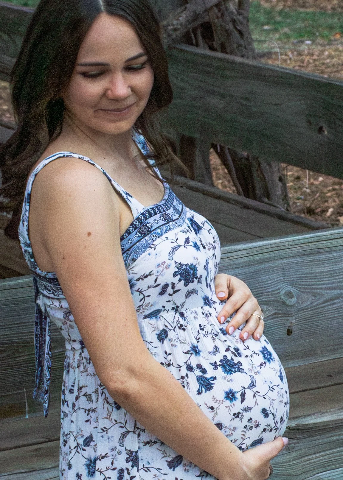 A pregnant woman in a sleeveless white dress with blue floral patterns, standing outdoors next to a wooden bench and tree. She is gently holding her stomach and has shoulder-length dark hair.