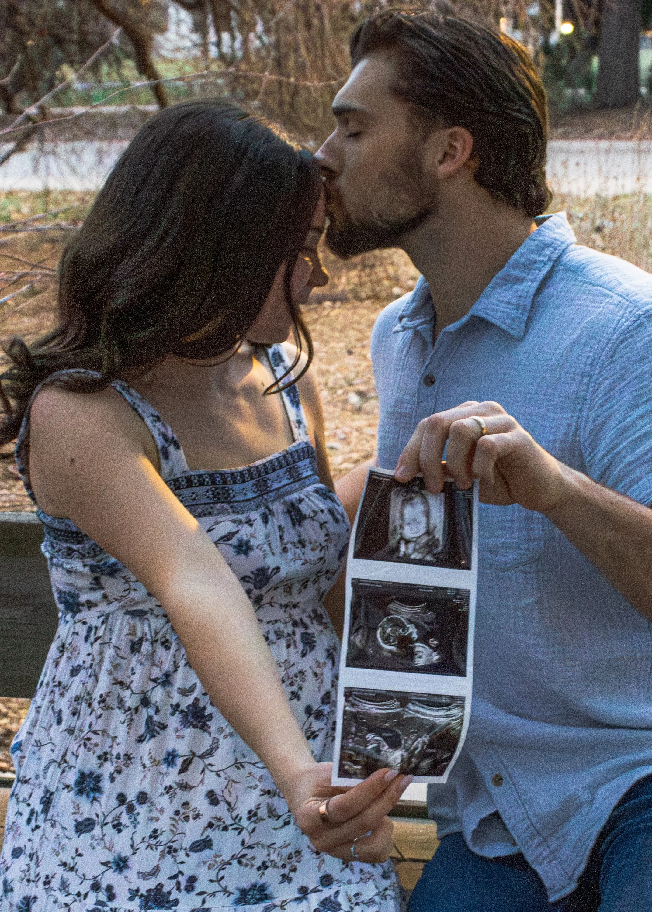 A man and woman are kissing in a park during sunset, holding ultrasound images of a baby.