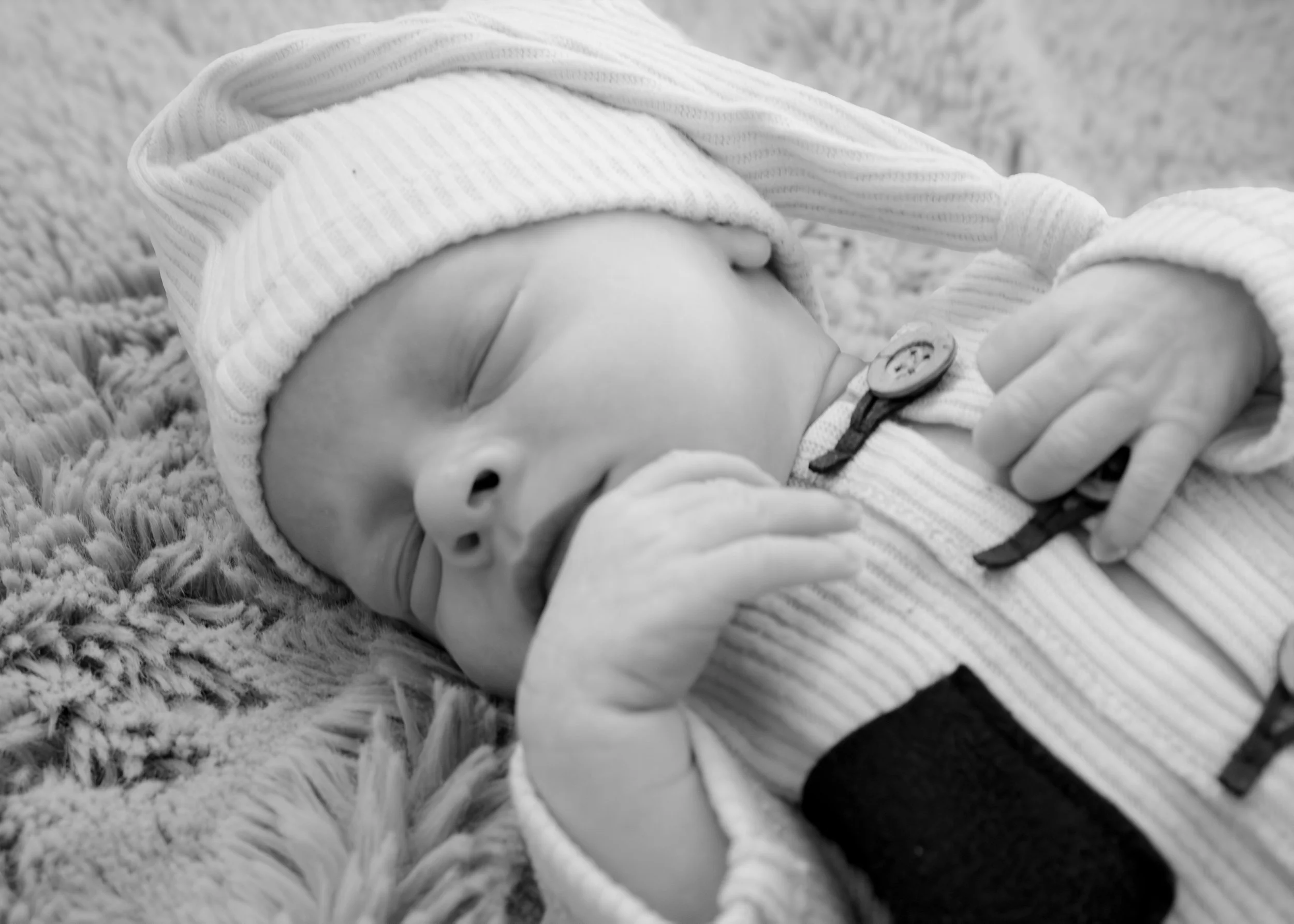 Close-up of a baby laying on a soft textured surface, wearing a striped hat and a onesie. The baby has a pacifier near their mouth and appears to be asleep or resting.