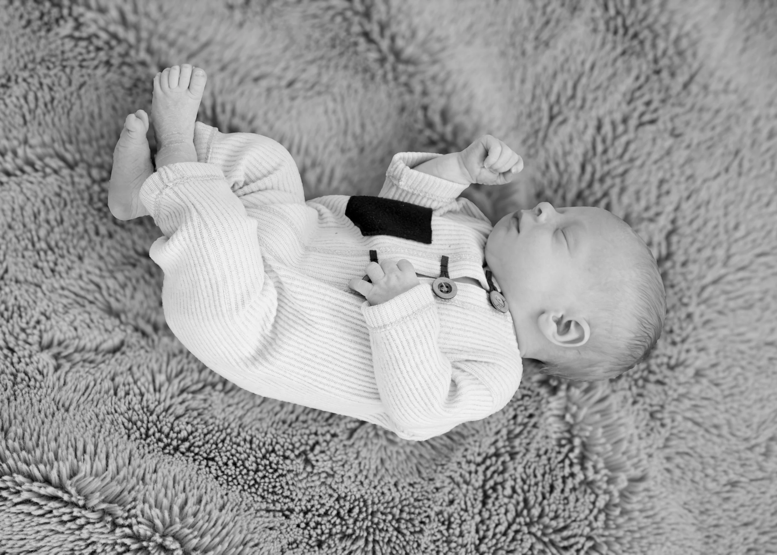 A baby lying on a fluffy textured blanket, wearing a striped sweater with buttons and a black collar. The baby appears to be relaxed with eyes closed and arms raised.