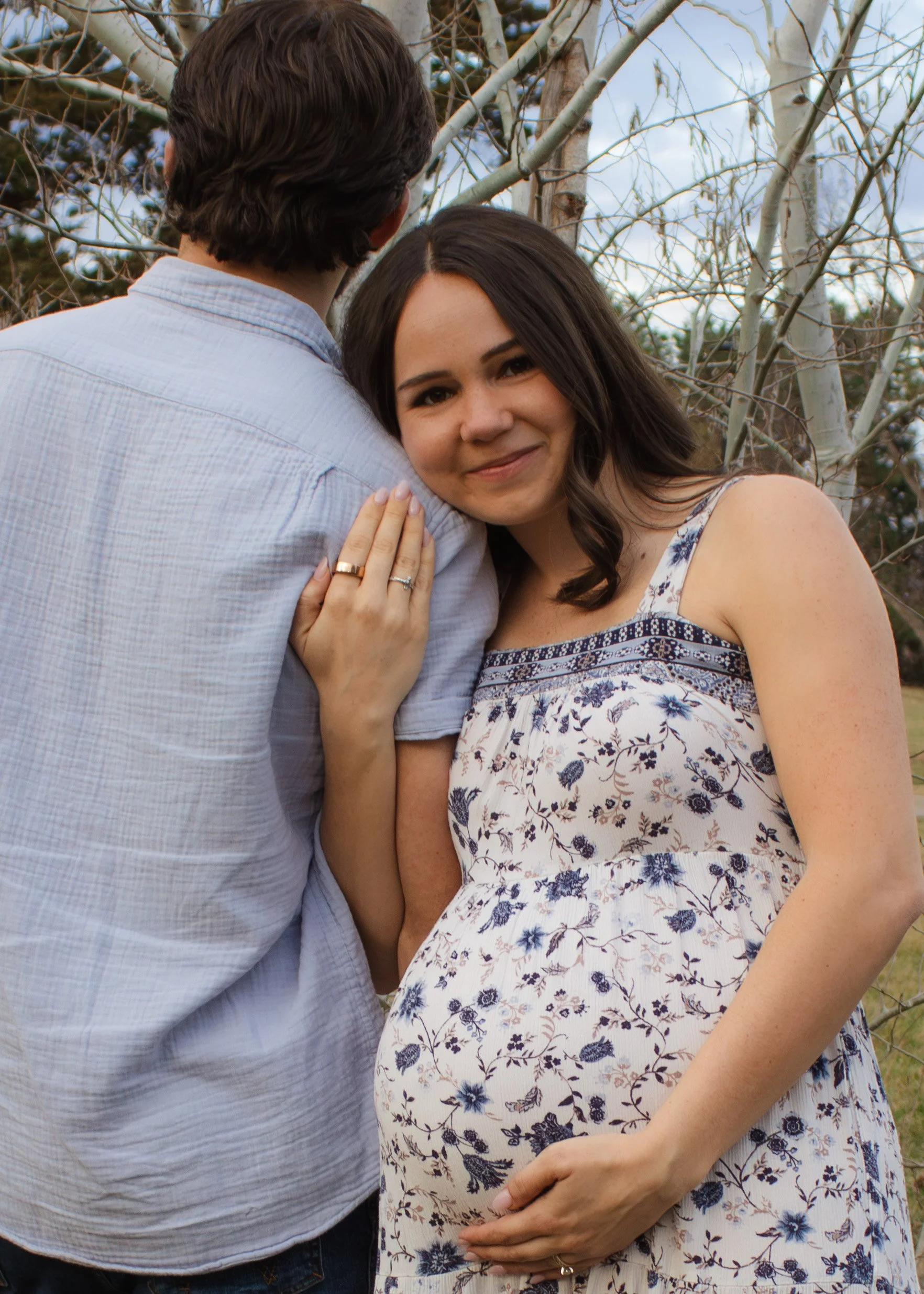 A pregnant woman with dark hair and a floral dress holds her belly, smiling, while her partner, who has dark hair, is standing close beside her, with his back turned. They are outdoors near leafless trees.
