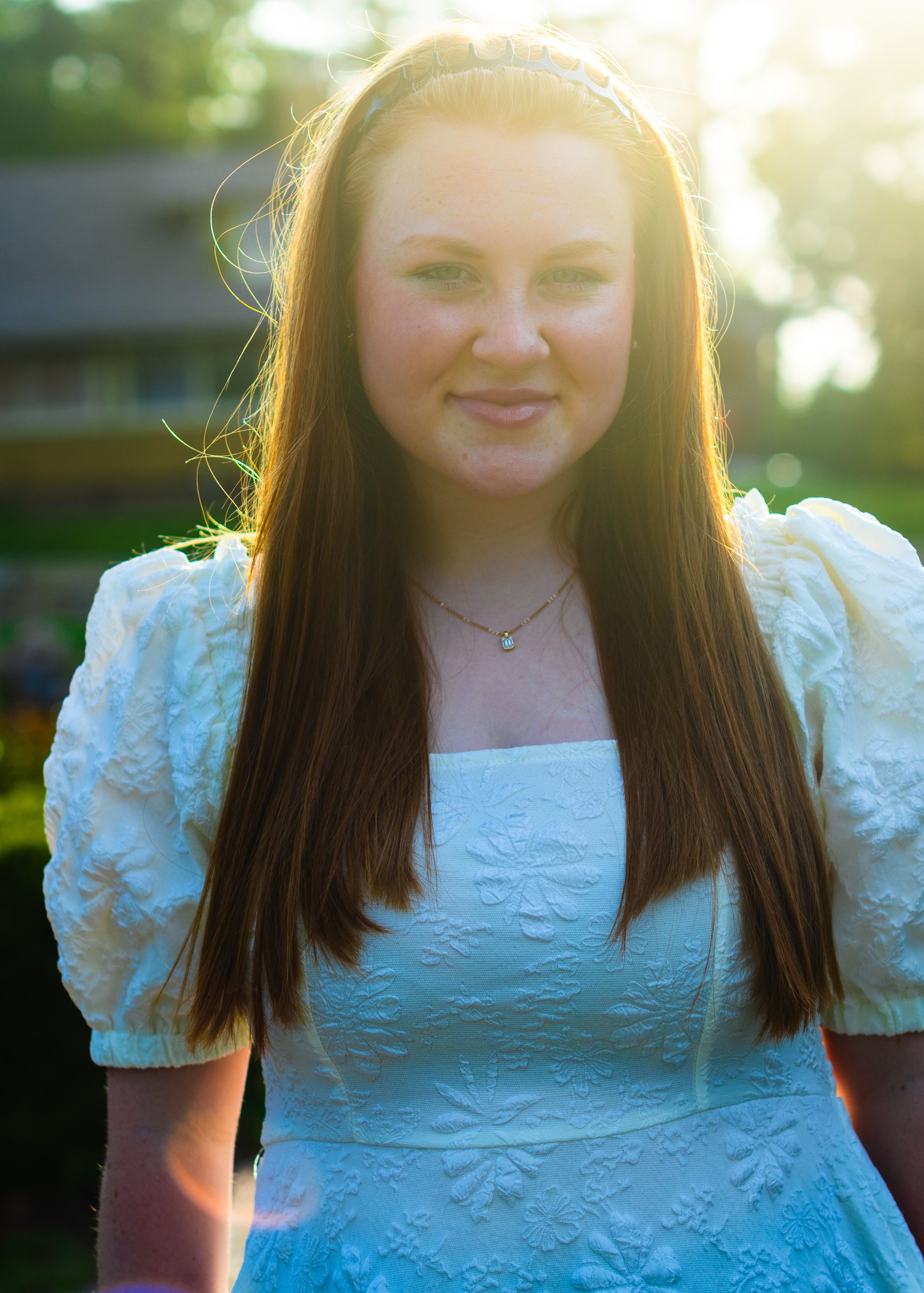 A young woman with long red hair, wearing a white dress with textured floral details and puffed sleeves, standing outdoors in sunlight.