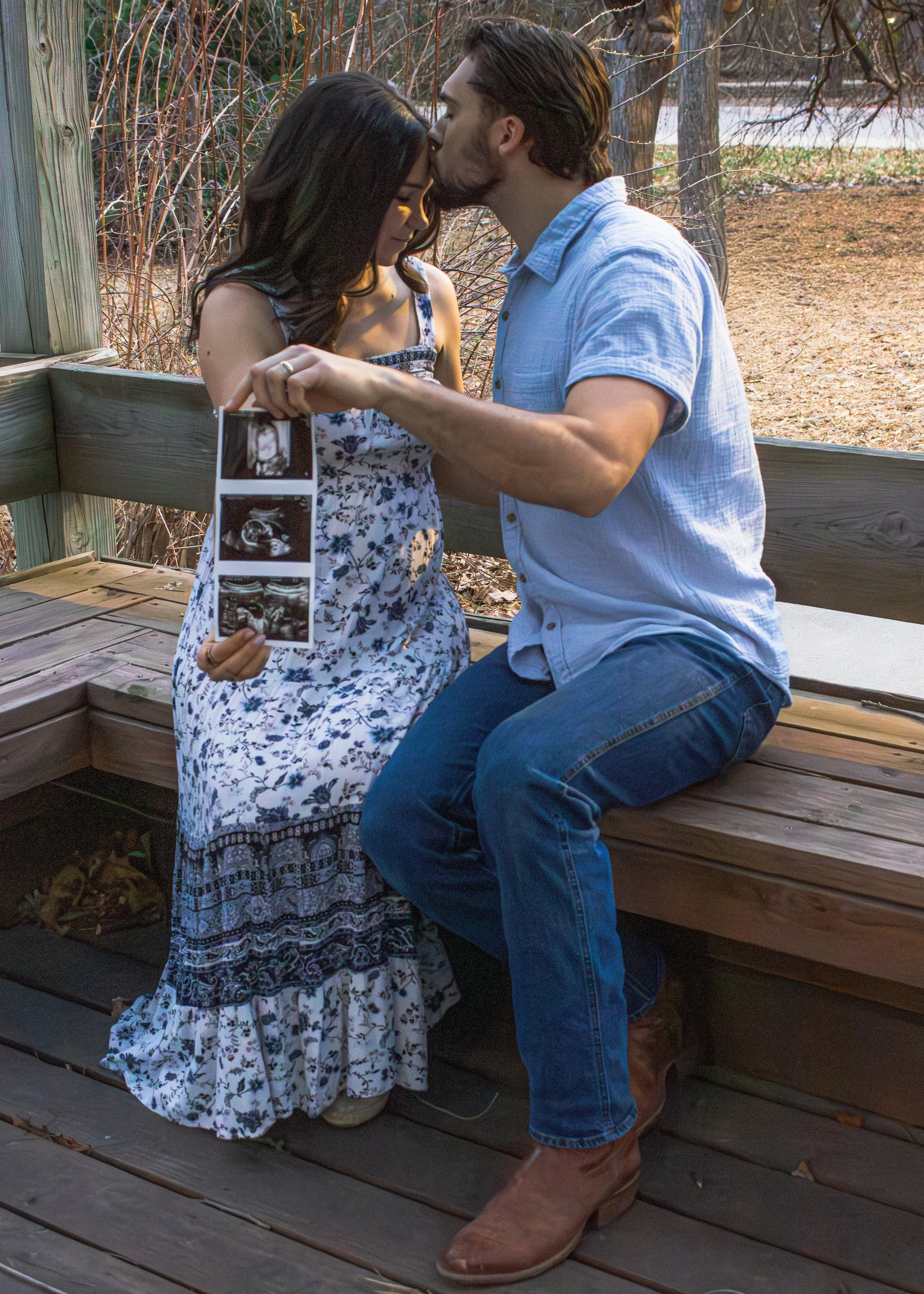 A man is kissing a woman on the forehead while holding her shoulders. The woman, dressed in a floral dress, is holding ultrasound images, indicating she is pregnant. They are outside on a wooden deck, with trees and a fence in the background.