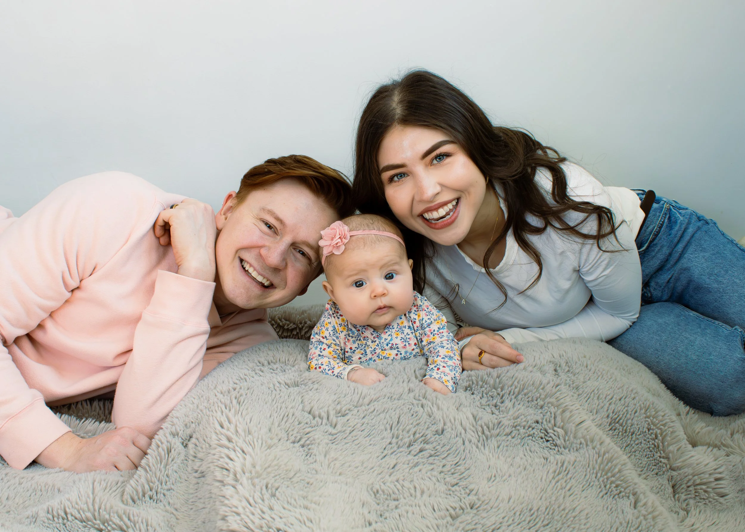 Four family members lying on a plush rug: a young man, a woman, a baby girl with a pink headband, and another baby girl with a floral dress, all smiling and close together.