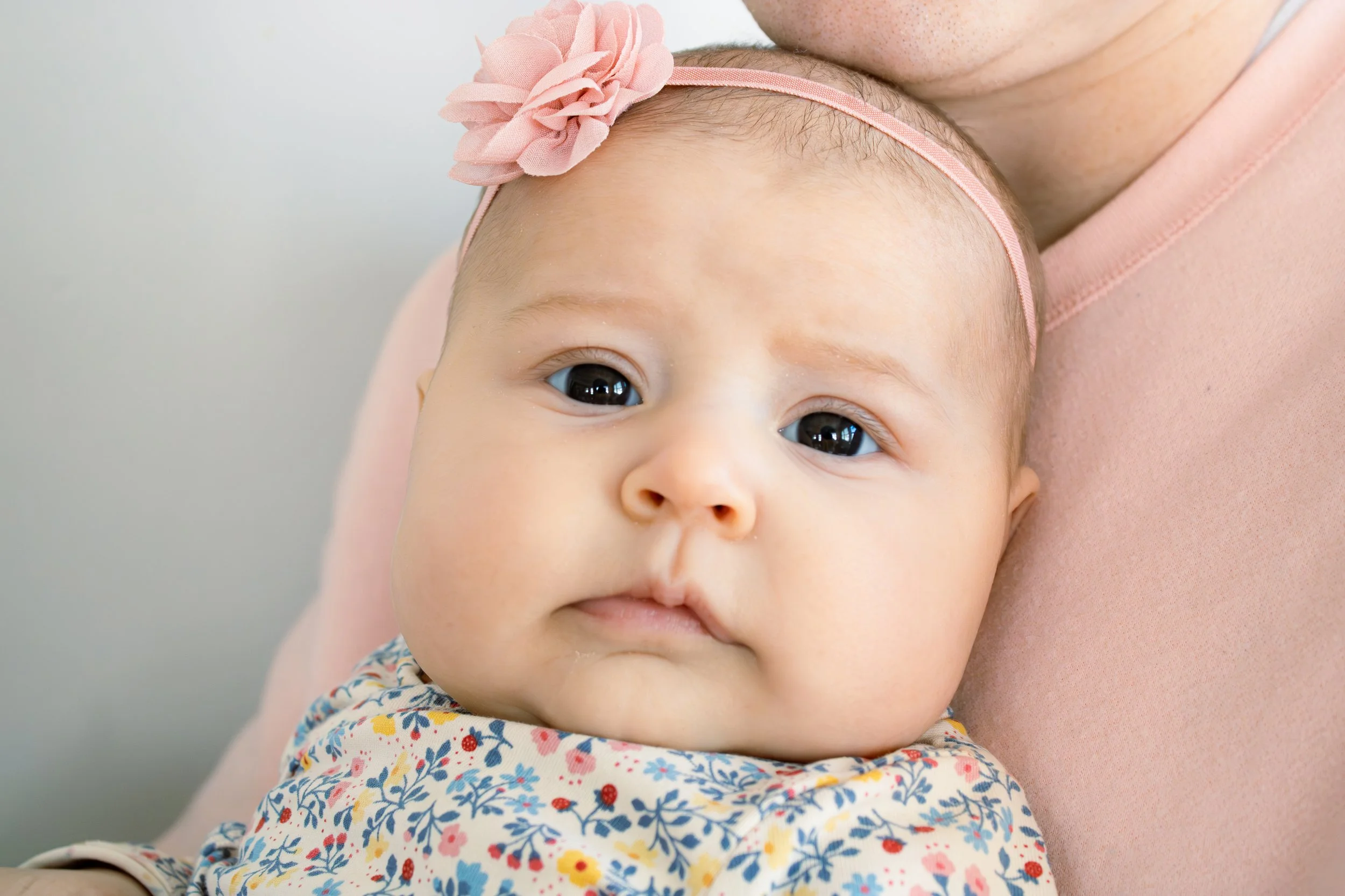 Close-up of a baby with big dark eyes, wearing a floral-patterned outfit, being held on someone's shoulder, with a pink headband with a flower. The person's arm is visible.
