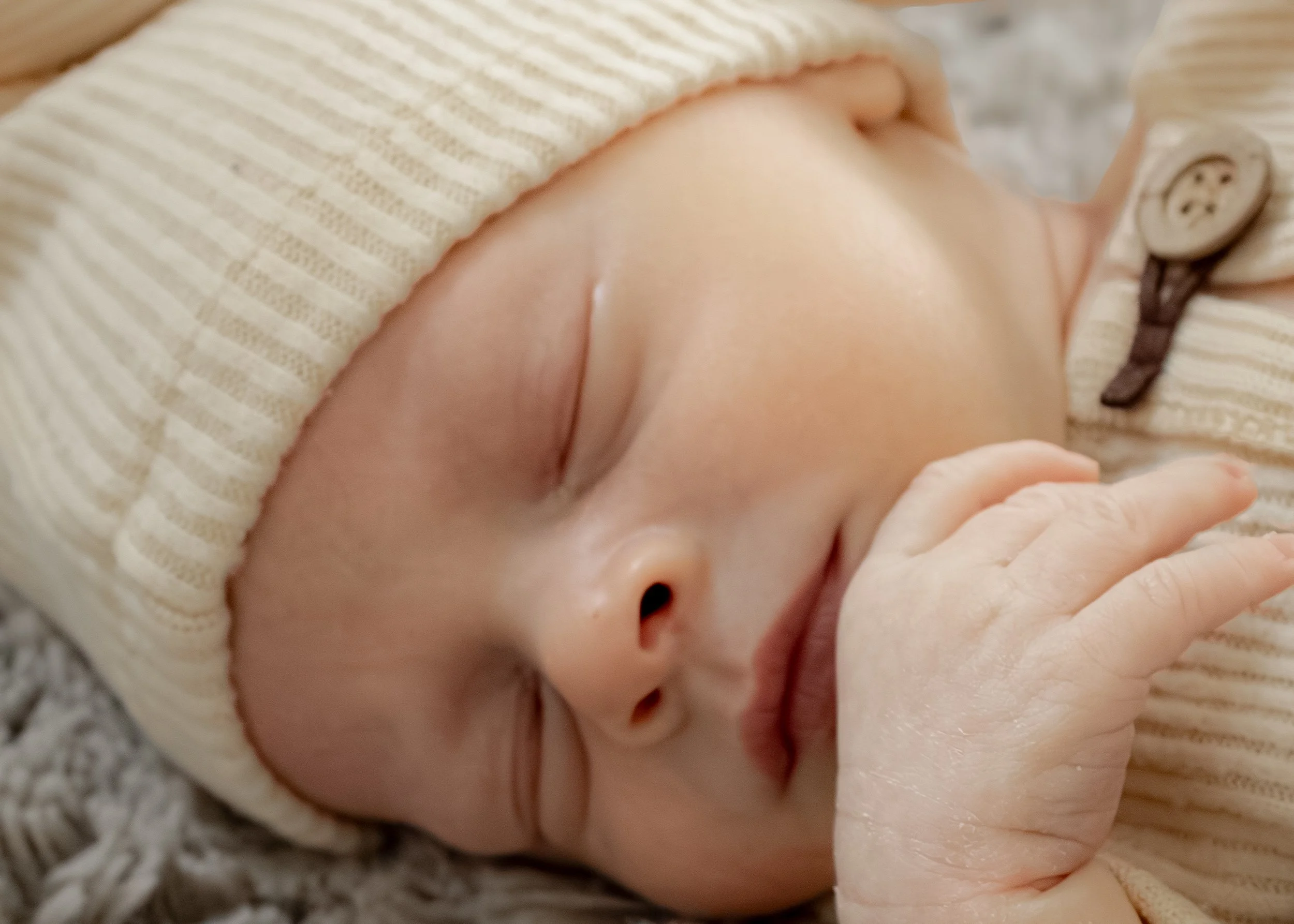Close-up of a newborn baby wearing a striped cream and brown beanie, lying on a soft surface, with a focus on the baby's head and tiny hand.