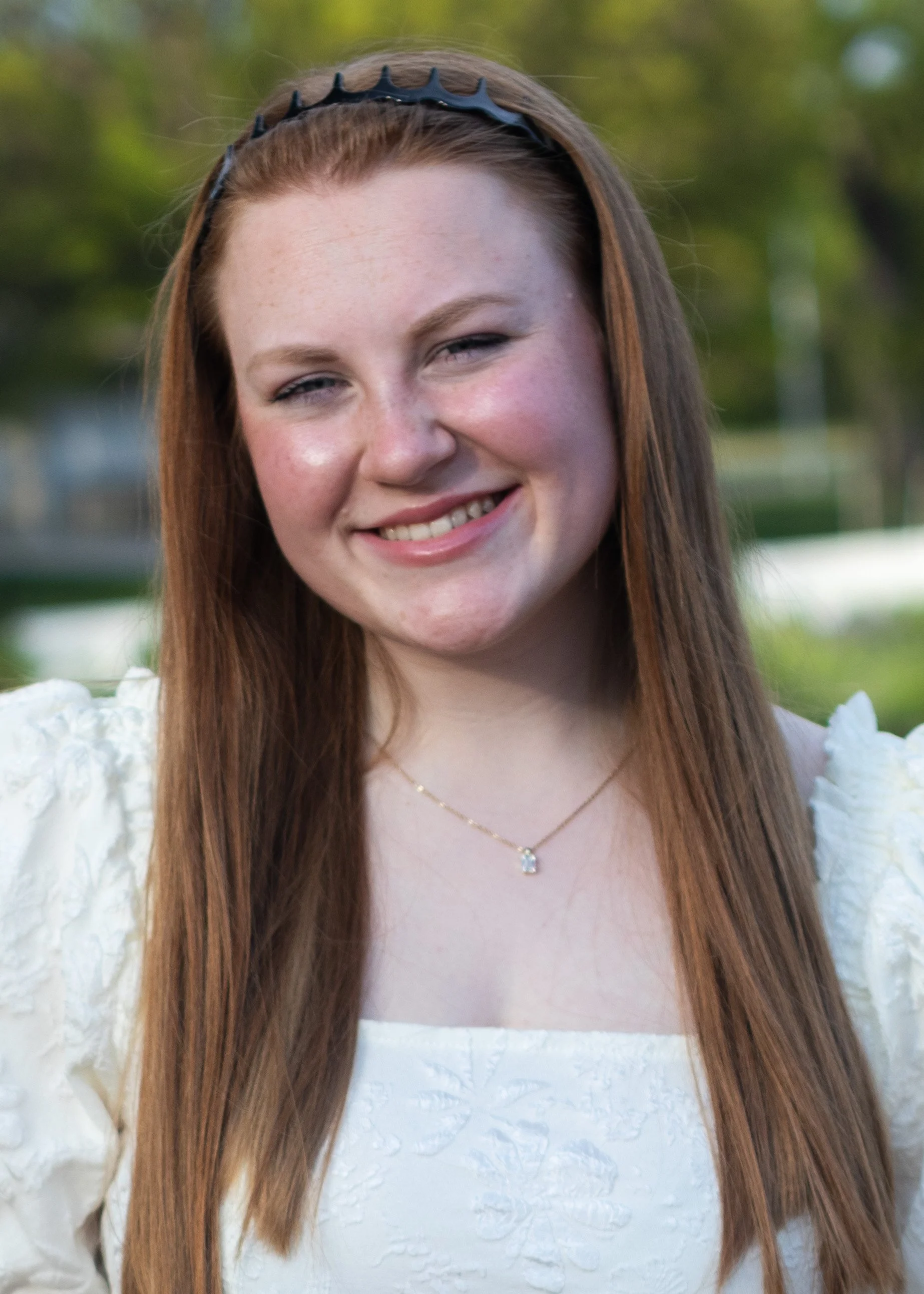 A young woman with long red hair, wearing a black hairband, a white dress with textured floral details, and a gold necklace with a small gemstone, smiling outdoors with blurred greenery in the background.